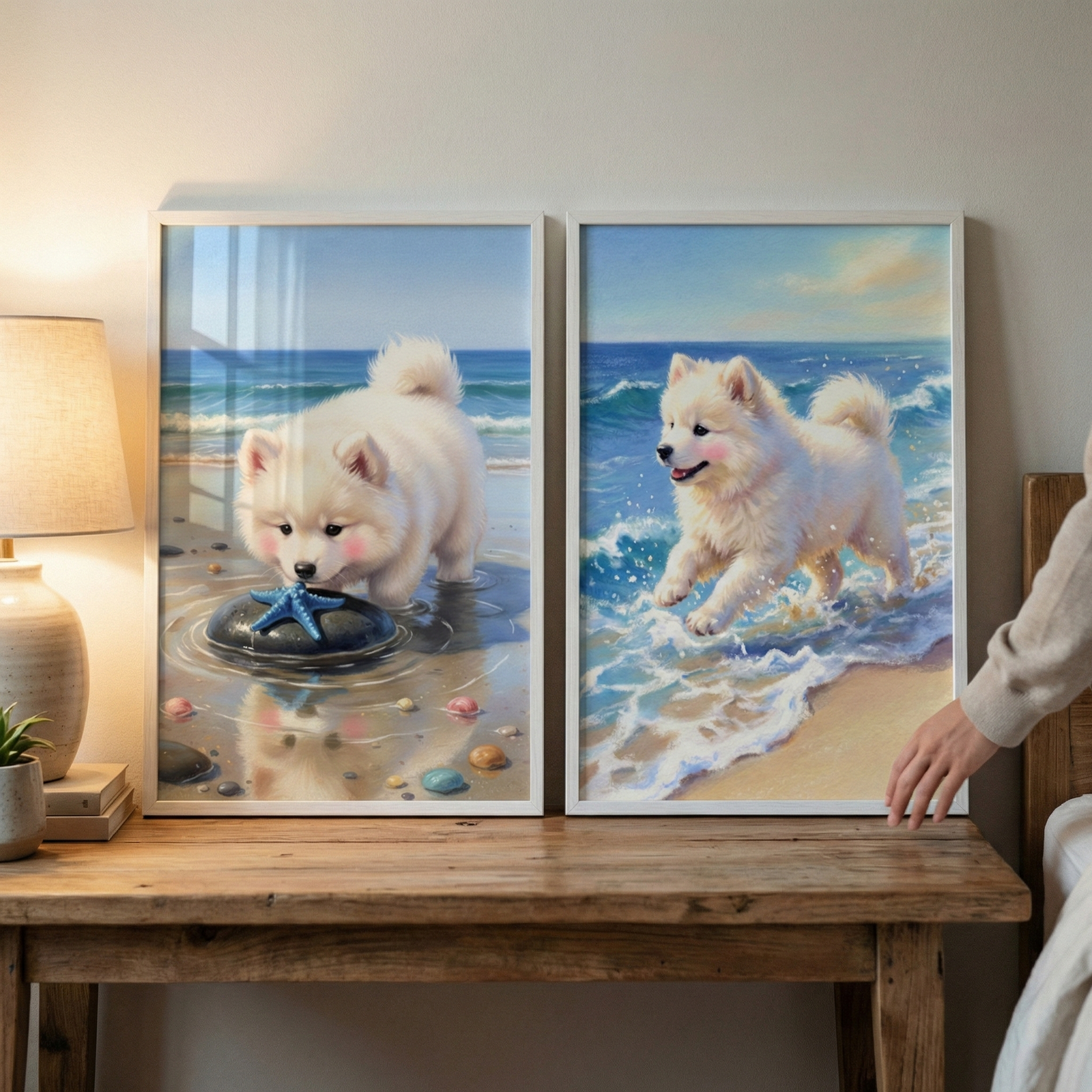 Two vertical white frames rest on a wooden table against a plain wall. Both frames contain art of a small, fluffy white dog at the seaside. The left piece shows the dog looking down at a blue starfish on a dark stone in the water. The right piece depicts the dog running through waves on a sandy beach. A lamp is on the left side of the table, and a person’s hand is visible on the right, touching the frame. The art is bright and has a soft, whimsical aesthetic.