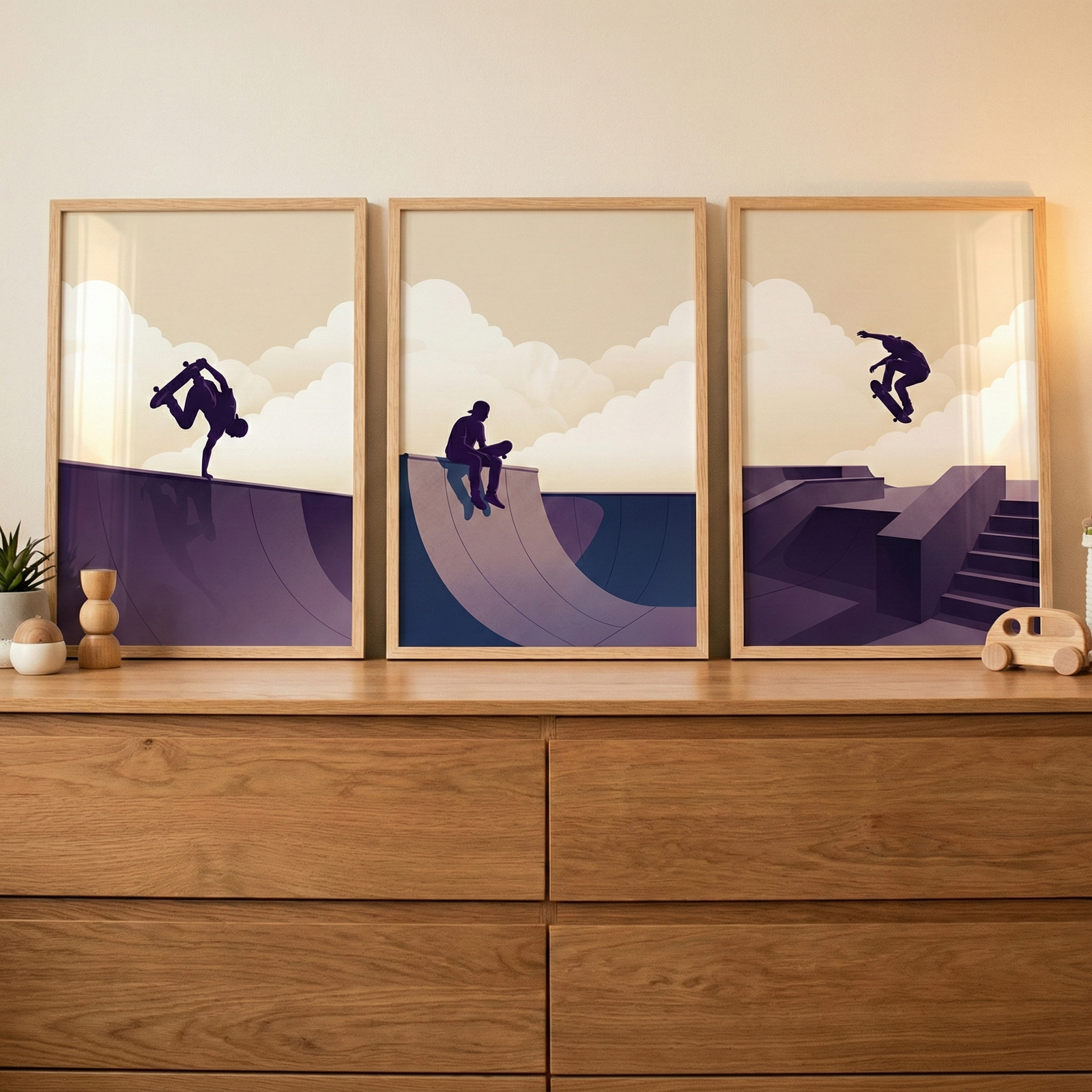 Three framed minimalist illustrations rest on a light wood dresser. The artwork features silhouettes of skateboarders in action against a backdrop of soft beige clouds and deep purple ramps. The left print shows a handplant on a ramp, the middle a person sitting on a ledge, and the right a jump over stairs. Beside the frames sit a small potted plant, a wooden decorative object, and a toy wooden car, all under warm indoor light.