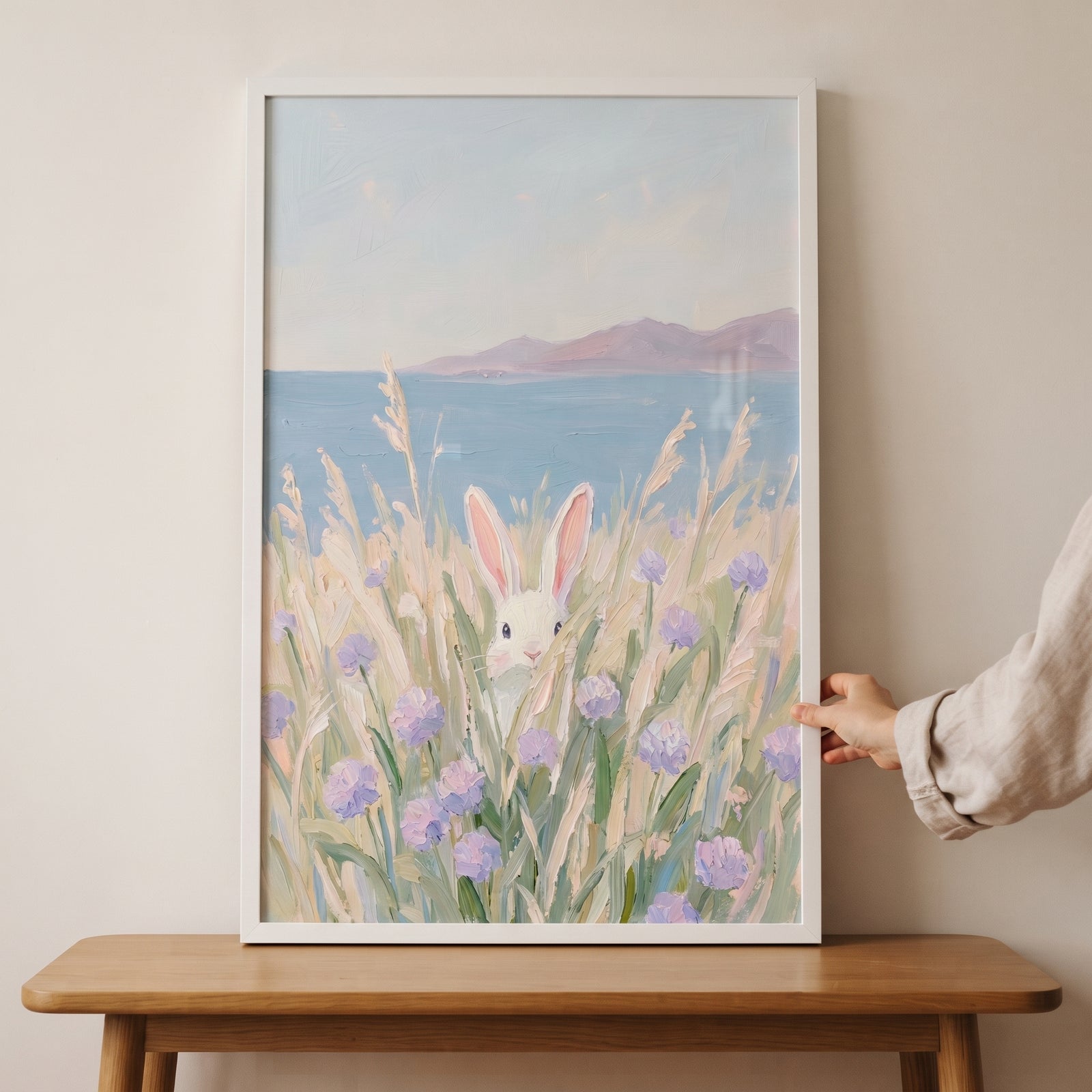 Purple wildflower peeking bunny wall art in a white frame, featuring a rabbit in a coastal field, held by a hand.