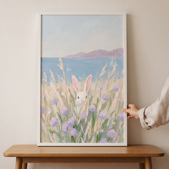 Purple wildflower peeking bunny wall art in a white frame, featuring a rabbit in a coastal field, held by a hand.