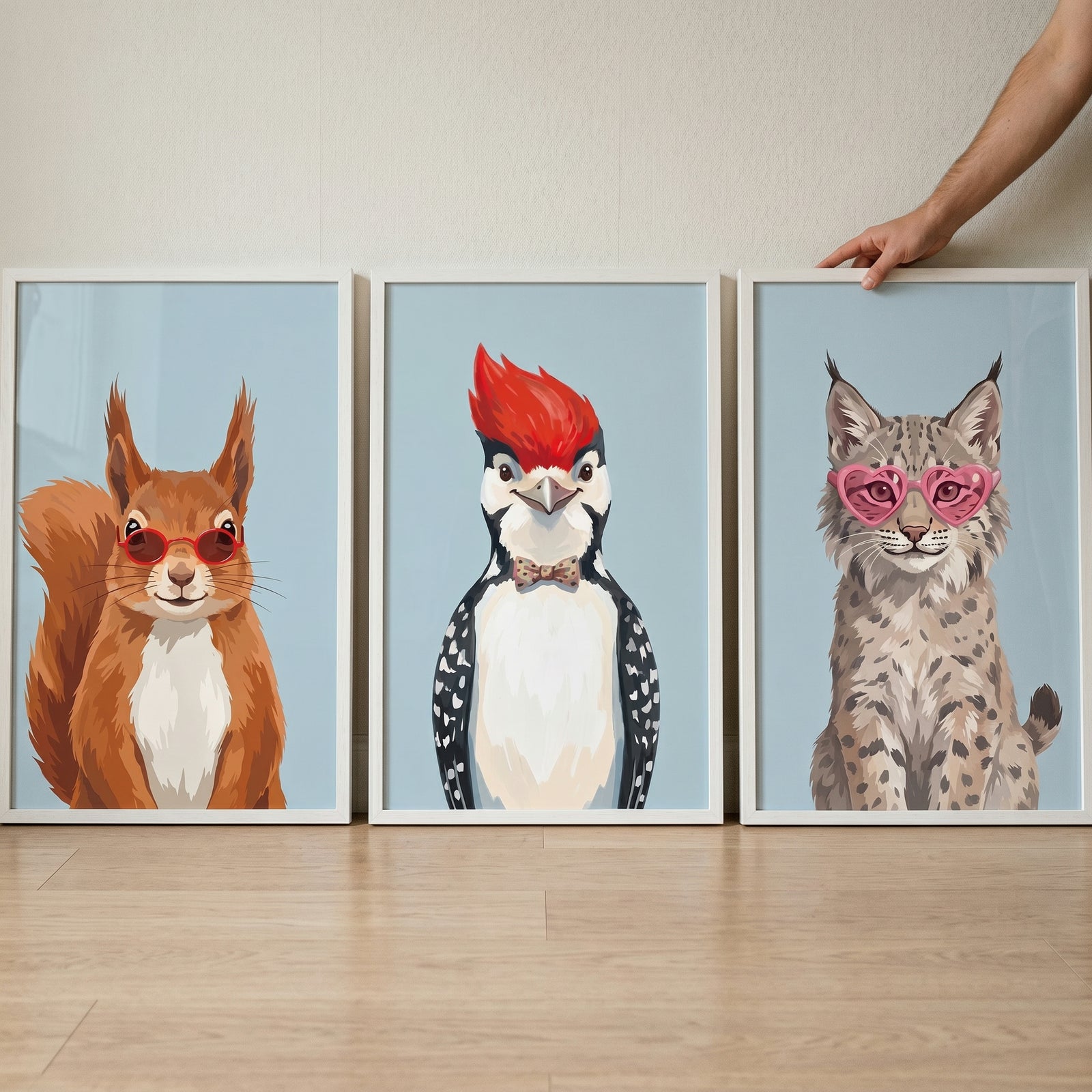 Three framed animal illustrations displayed side by side featuring a squirrel wearing red sunglasses, a woodpecker with a bright red crest and bow tie, and a lynx wearing pink heart shaped sunglasses. A hand adjusts the lynx frame, creating a playful gallery style wall art display.