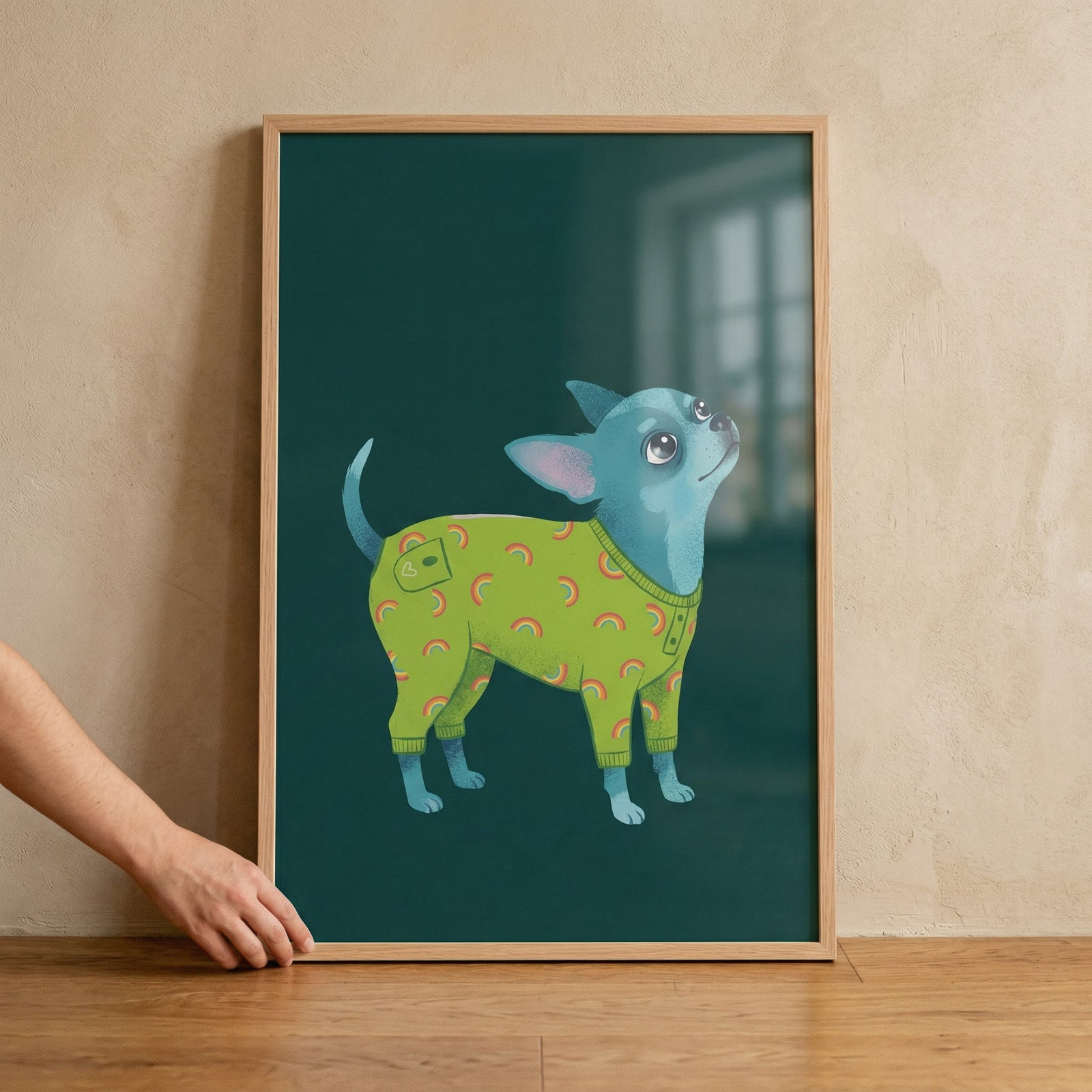 A framed digital illustration features a small, blue-colored dog looking upwards with large, expressive eyes. The dog is dressed in bright green pajamas with a pattern of small rainbows. The background of the artwork is a solid, dark teal. 