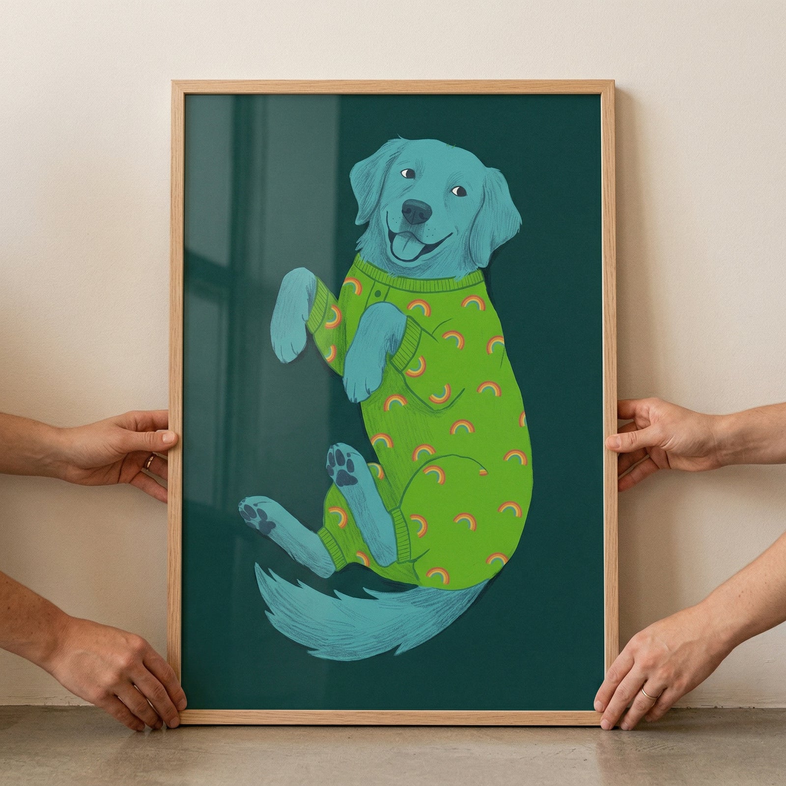 A framed piece of artwork is held up against a plain, light-colored wall by two sets of hands. The illustration depicts a light blue dog in a playful, belly-up position with a cheerful expression. The dog is wearing bright green pajamas adorned with small orange rainbow patterns. 