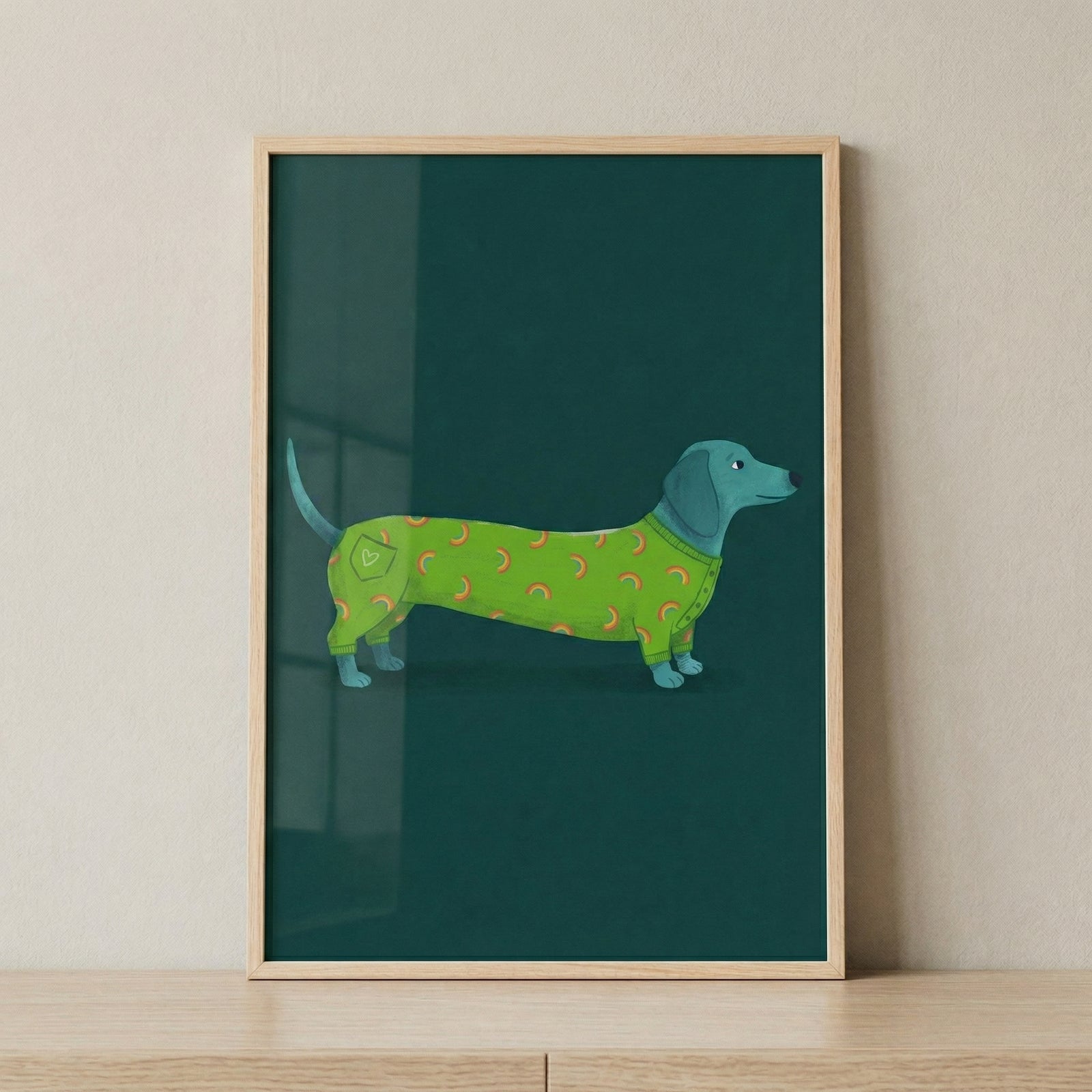 A minimalist, flat-style digital illustration of a long-bodied, teal-blue dog with short legs. The dog is positioned horizontally and is wearing a bright green, full-length garment that covers its entire torso. The outfit is patterned with small, scattered orange crescent moons and features a small pocket on the back hip with a heart design. 