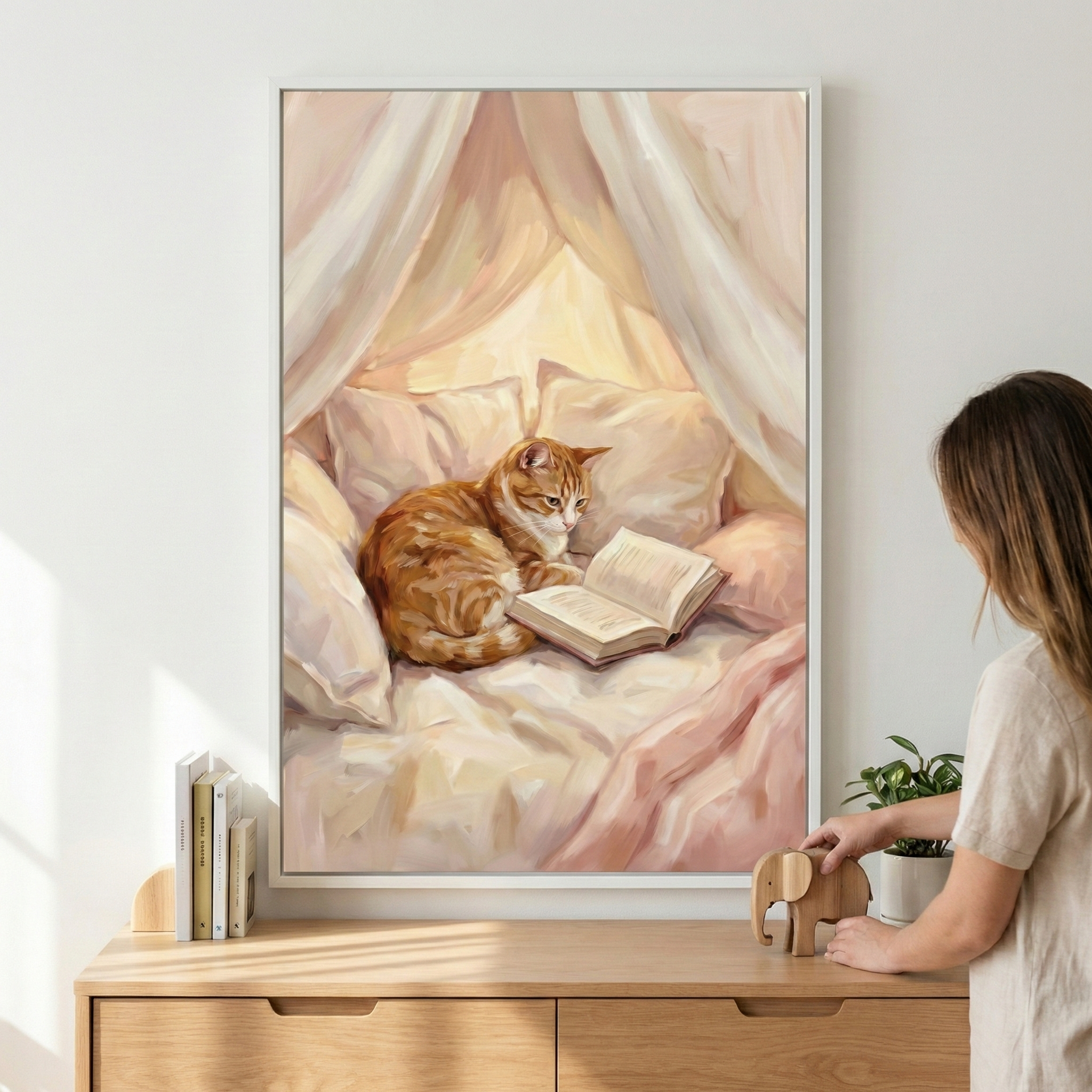 A framed vertical artwork captures a cozy scene of an orange tabby cat resting on a plush bed, seemingly engrossed in an open book. The cat is nestled among soft pillows beneath a draped canopy of sheer, light-colored fabric that glows with a warm, golden radiance. The style uses soft brushstrokes and a gentle palette of peach and cream. The piece is displayed in a white frame on a wall above a wooden dresser, where a hand reaches toward a small wooden elephant figurine.