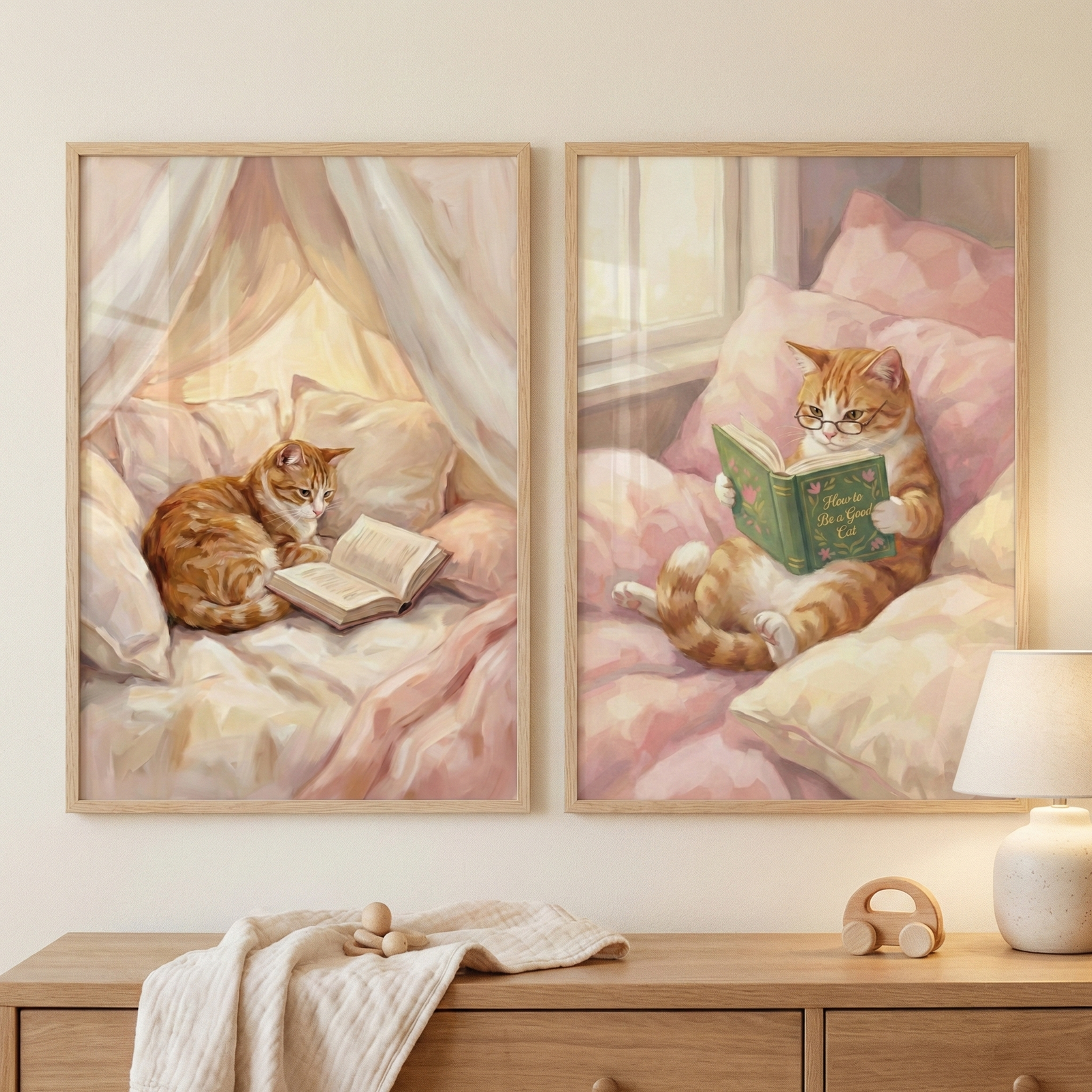 Two framed illustrations hang side-by-side on a neutral-toned wall above a light wood dresser. Both pieces depict a ginger and white tabby cat in cozy scenes. The left artwork shows the cat curled up on a soft bed under a canopy, looking at an open book. The right artwork features the cat sitting upright against pink pillows, wearing spectacles while reading a green book. The style is soft and illustrative with a pastel palette. A wooden toy car and a lamp sit on the dresser below.