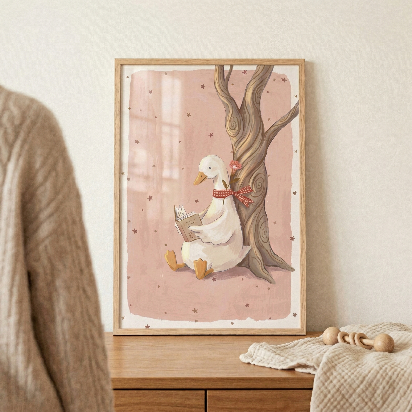 A framed illustration sits on a light wooden surface, depicting a whimsical and gentle scene. In the center of the artwork, a white duck with orange feet and a beak is sitting comfortably against the base of a tall, winding tree trunk. The duck is holding an open book and appears to be reading. A red and white checkered ribbon is tied around its neck, with a small pink blossom tucked into the bow.