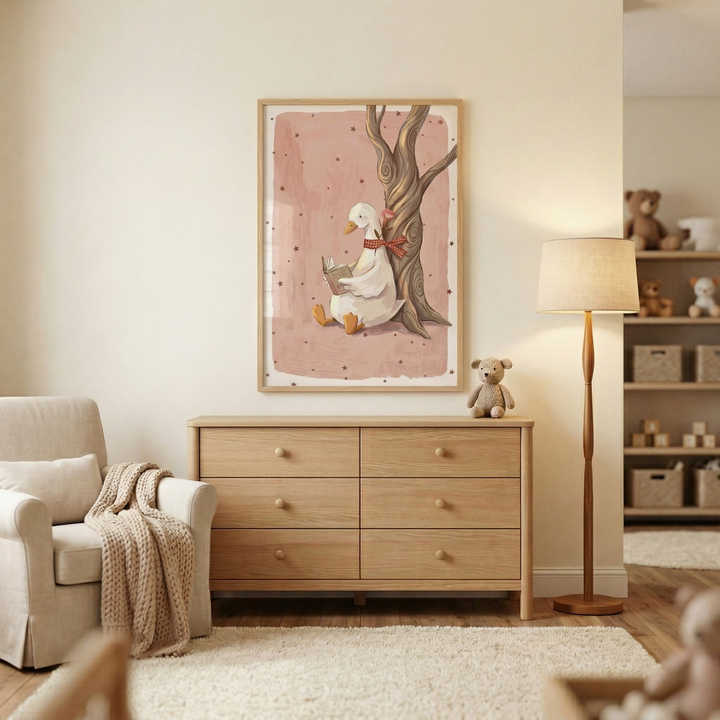 A cozy nursery featuring a large framed illustration of a white goose with a red bow reading a book. The art is set against a pink background and hangs above a light wood six-drawer dresser. To the left is a cream armchair with a textured throw blanket. A tall wooden floor lamp with a beige shade stands nearby, and a small teddy bear sits on the dresser. The room has neutral walls, a white rug, and a wooden bookshelf with toys in the background, creating a calm, minimalist atmosphere.