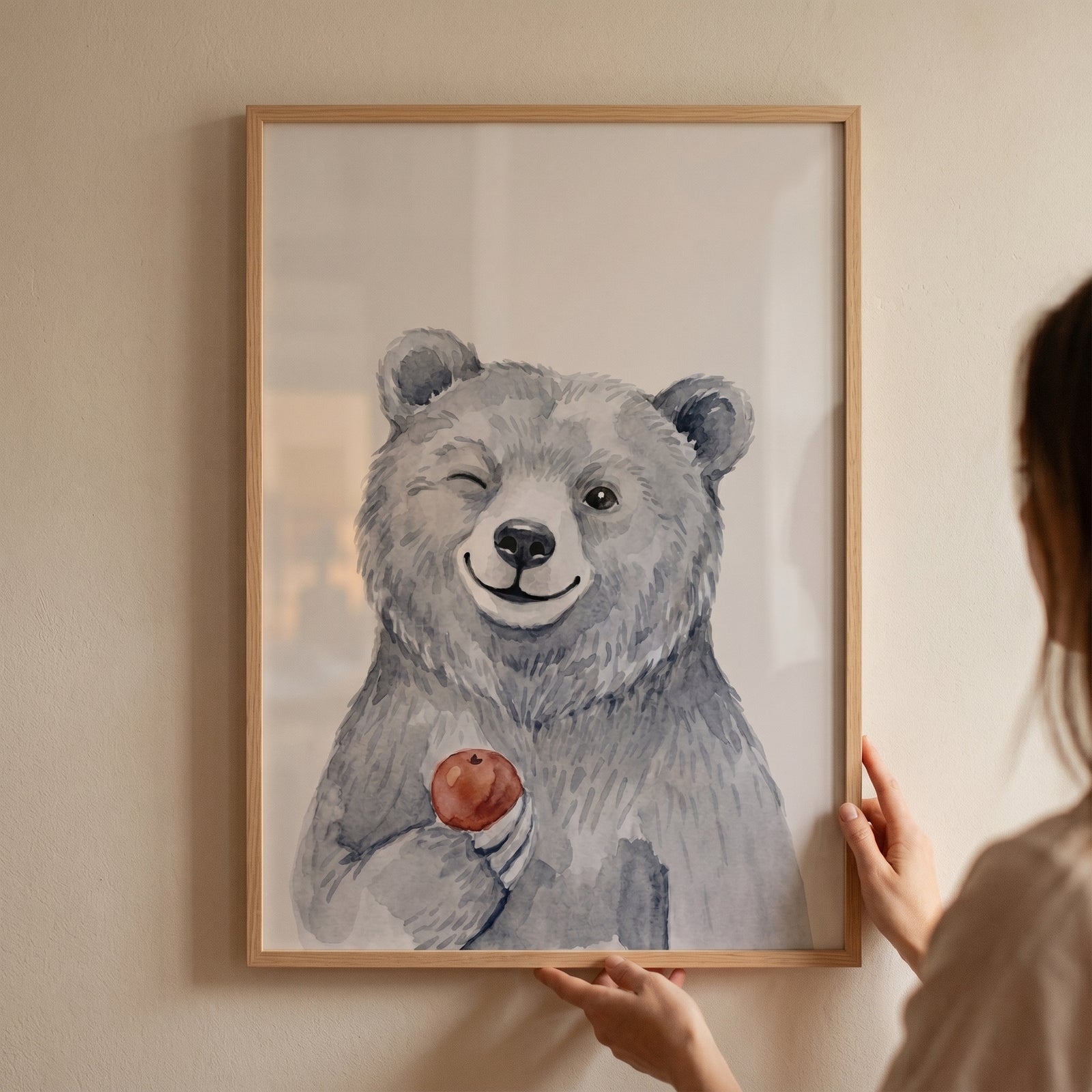 Hands hold a framed red apple winking bear artwork showing a smiling watercolor bear on a neutral wall.