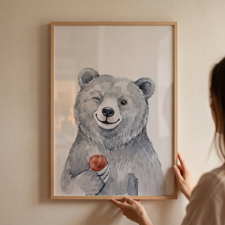 Hands hold a framed red apple winking bear artwork showing a smiling watercolor bear on a neutral wall.