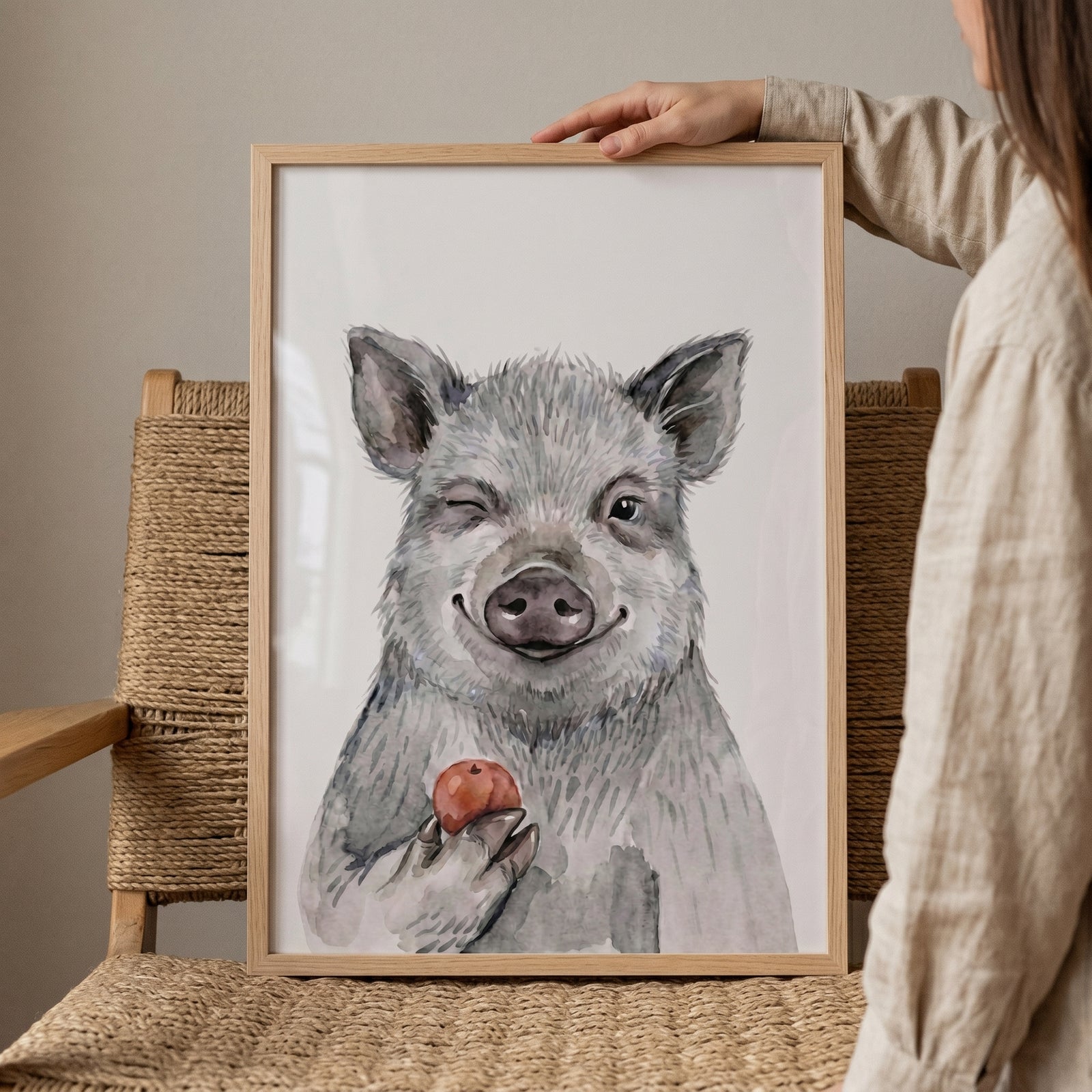 Framed red apple winking pig wall art resting on a woven chair, held by a person against a neutral wall.