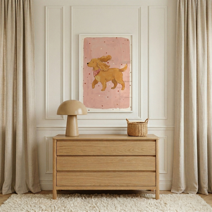 A minimalist interior scene features a light-colored wooden three-drawer chest centered against a white paneled wall. Hanging above the chest is a framed illustration of a cheerful, golden-brown dog with long, floppy ears wearing a red and white checkered bandana. The dog is depicted mid-stride against a soft pink background dotted with subtle specks. 
