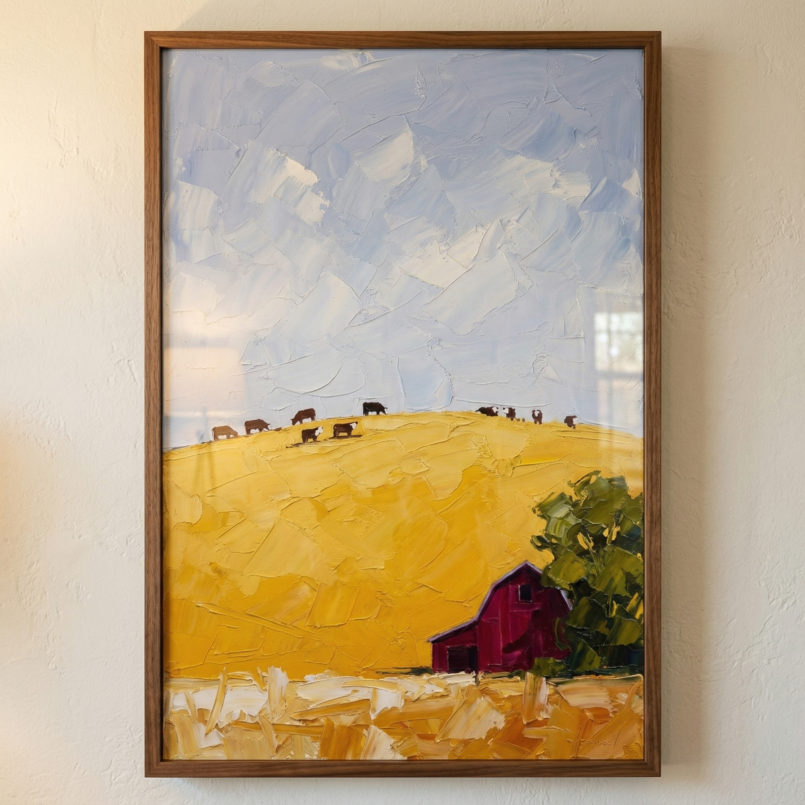 A vertical framed artwork hangs on a light-colored wall. The scene depicts a vibrant rural landscape rendered with thick, textured brushstrokes. A vast, pale blue sky with white clouds fills the top half. Below, a sloping golden-yellow hill is dotted with a small herd of cows along its crest. In the lower right, a deep red structure sits beside a lush green tree. The bottom edge of the artwork features a field of light gold and cream, showing prominent texture and depth. The entire piece is encased in a sim
