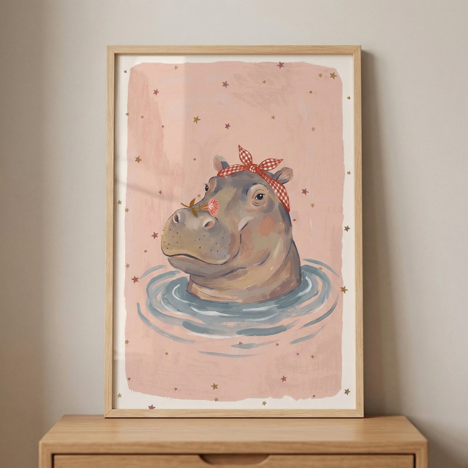 Framed illustration of a hippo floating in water with gentle ripples, wearing a red checkered headband and holding a small flower near its nose against a soft pink background with scattered stars. The playful artwork adds a calm and whimsical touch to a nursery or children’s room decor.