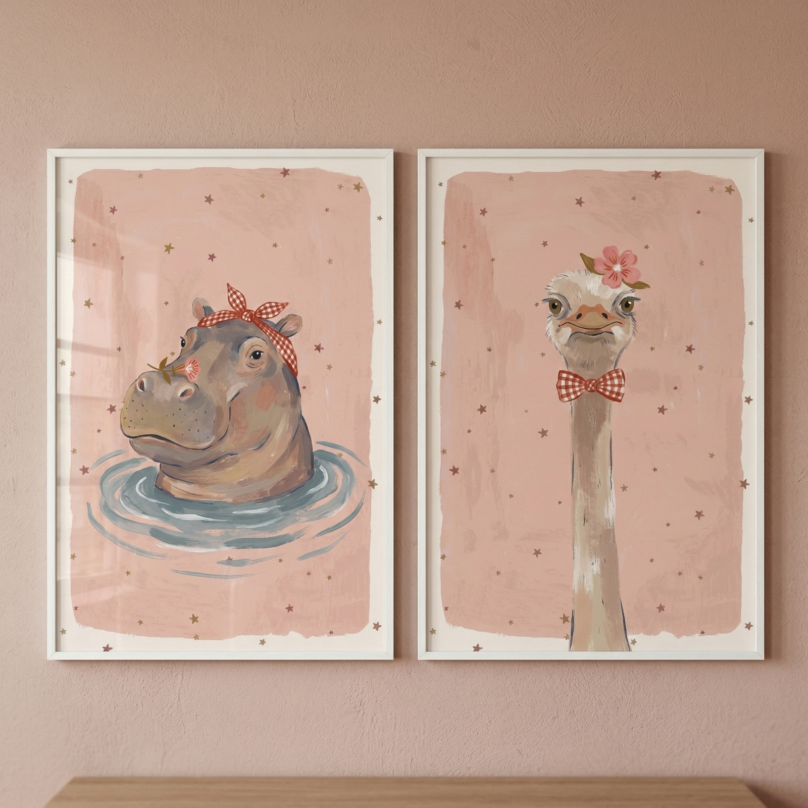 Two side-by-side, white-framed vertical illustrations hang on a dusty rose-colored wall. The left illustration depicts a whimsical hippopotamus with its head above blue water ripples, wearing a red and white checkered bow. The right illustration shows the long neck and head of an ostrich wearing a small pink flower and a matching red and white checkered bow tie. Both artworks have a soft, textured pink background decorated with small, scattered brown stars.