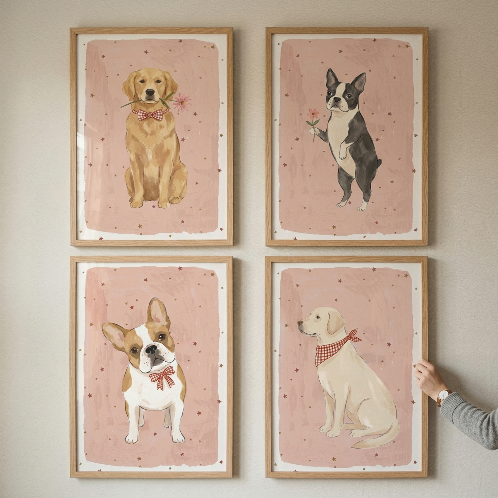 Four framed art prints are arranged in a two-by-two grid on a light-colored wall. Each illustration features a different dog against a soft pink, textured background with tiny star accents. The top left shows a golden dog holding a flower and wearing a checkered bowtie. The top right depicts a black and white dog standing on its hind legs with a flower.