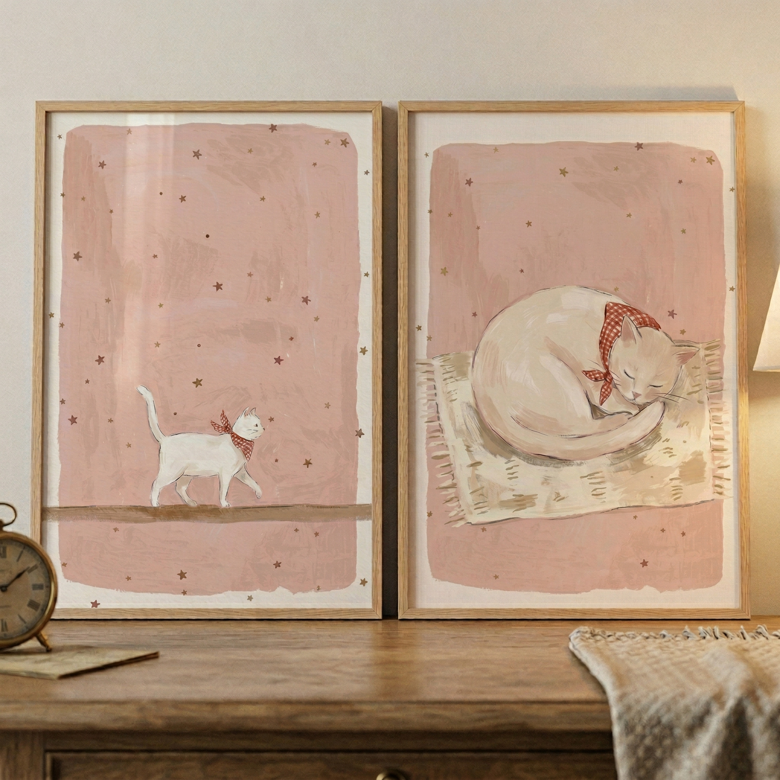 Two light wood-framed illustrations sit on a wooden surface against a neutral wall. The left image shows a small white cat wearing a red and white checkered neckerchief, walking along a thin line. The background is a soft pink decorated with tiny stars.
