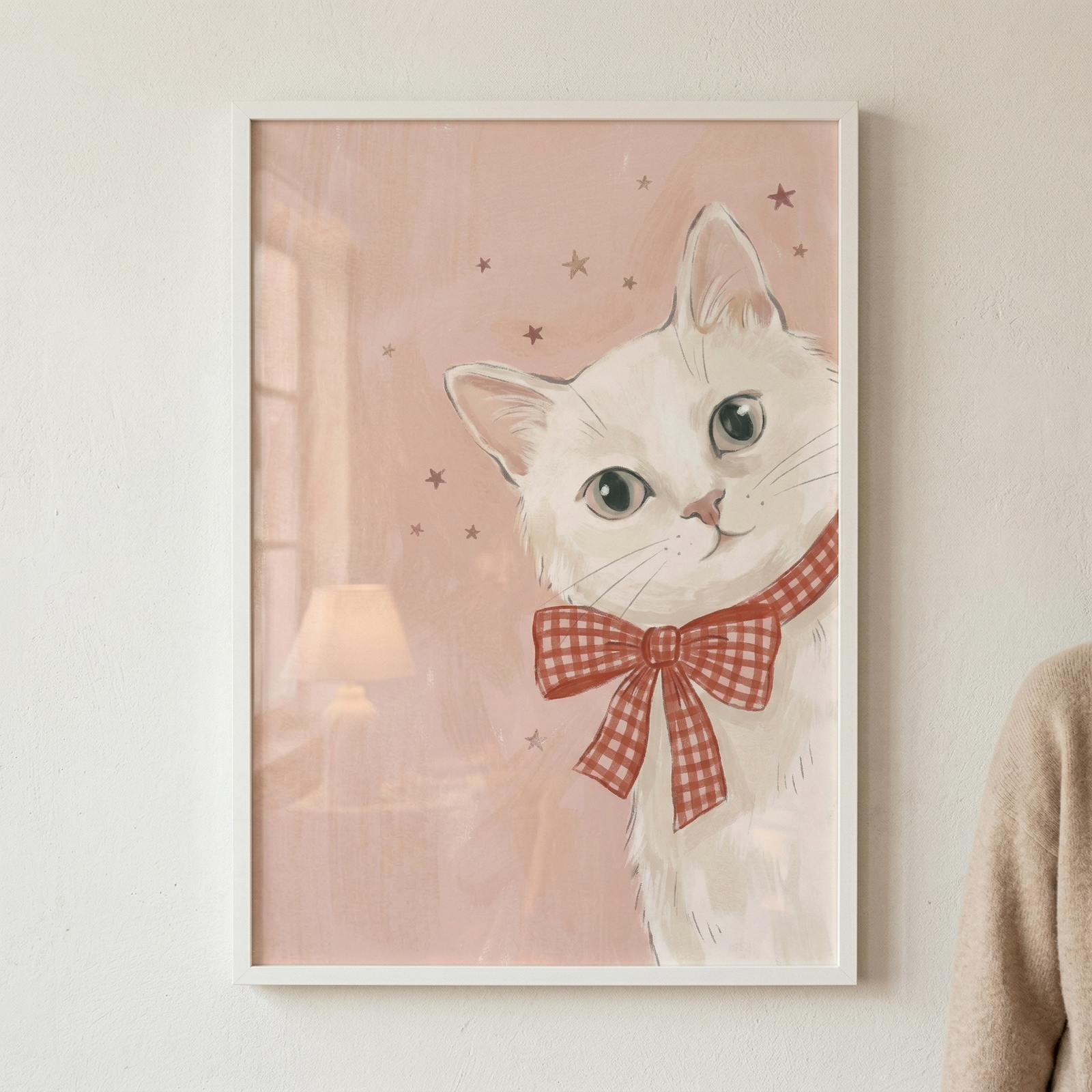 A framed vertical illustration of a white cat with large, light-colored eyes looking toward the viewer. The cat wears a large red and white checkered bow around its neck. The background is a soft, muted pink decorated with a few small, dark stars. In the blurry background on the left, there is a faint suggestion of a window and a lamp. The artwork is in a thin white frame, hanging on a light, textured wall. Part of a person wearing a beige sweater is visible on the far right.