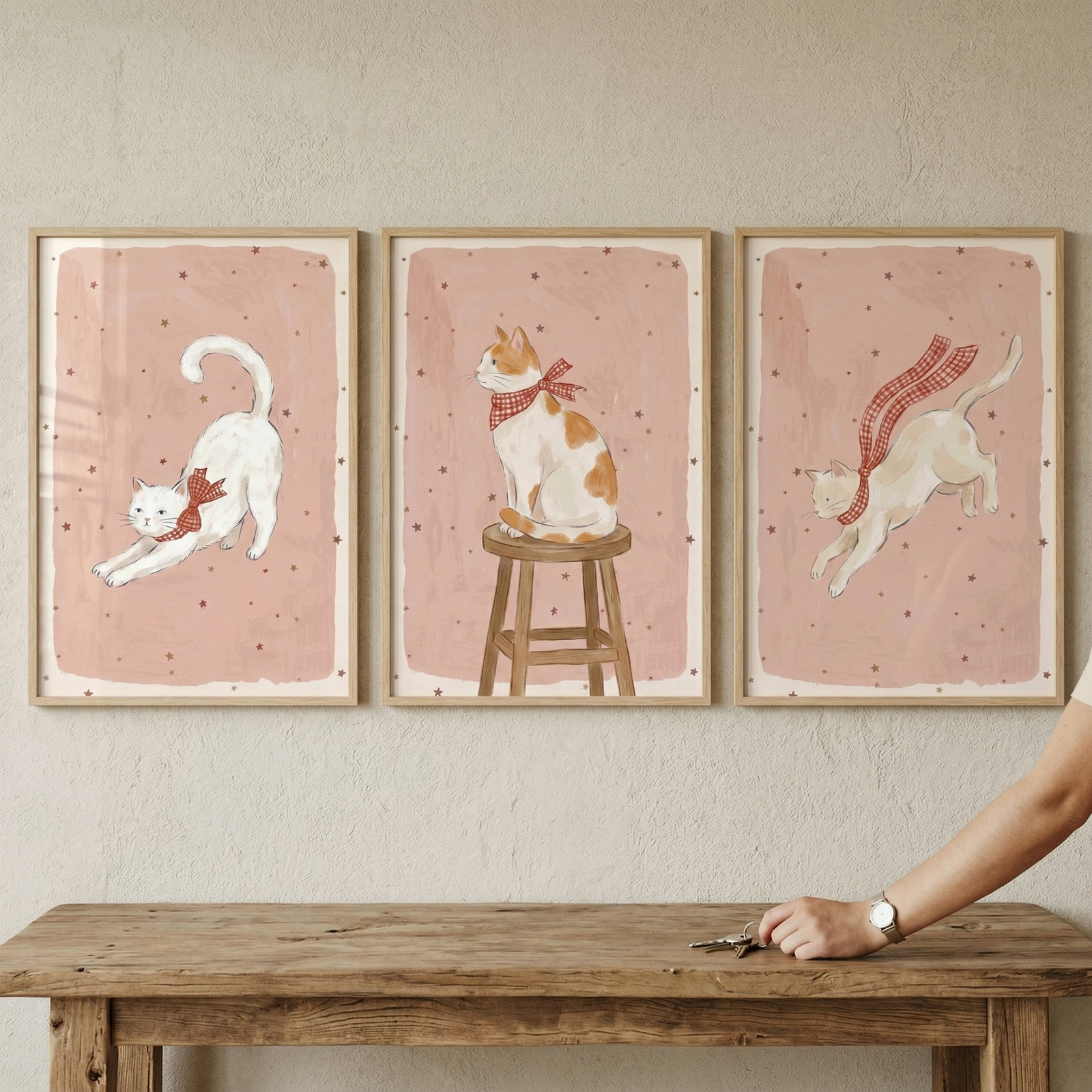 A set of three light wood-framed art prints hangs on a textured wall. Each piece features a charming illustration of a cat set against a soft pink background with a pattern of small stars. From left to right, the first shows a white cat stretching while wearing a red checkered bow. 