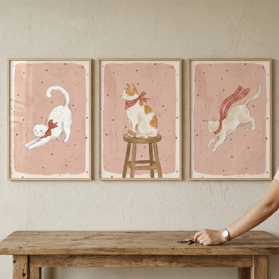 A set of three light wood-framed art prints hangs on a textured wall. Each piece features a charming illustration of a cat set against a soft pink background with a pattern of small stars. From left to right, the first shows a white cat stretching while wearing a red checkered bow. 