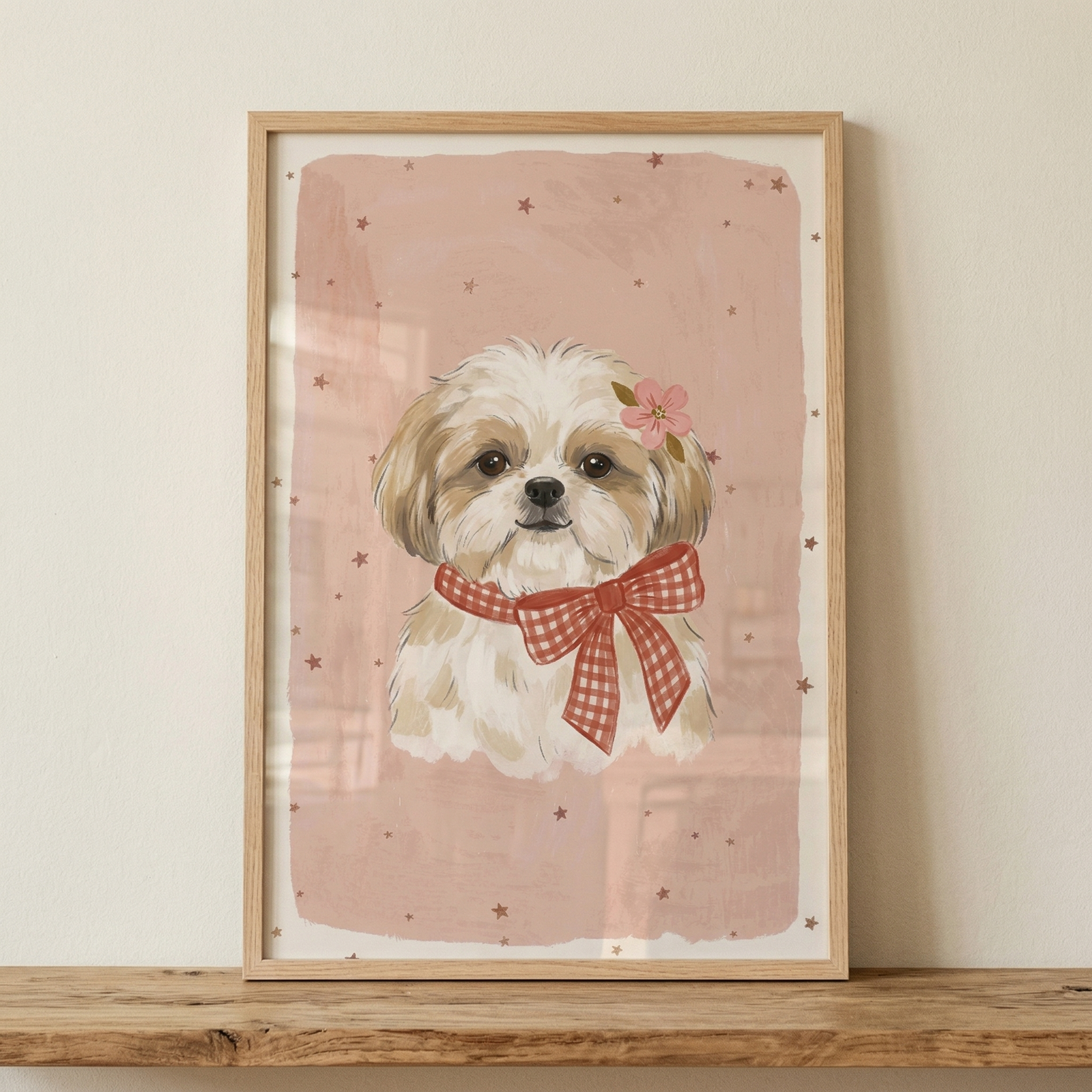 A framed illustration of a small, fluffy dog sits on a rustic wooden shelf. The dog has white and tan fur, expressive dark eyes, and a black nose. It is adorned with a large red and white checkered bow around its neck and a delicate pink flower tucked by its ear. The background of the artwork is a textured dusty pink with small dark stars and is bordered by a white frame. The piece is held in a light-colored wooden frame, leaning against a neutral-toned wall.