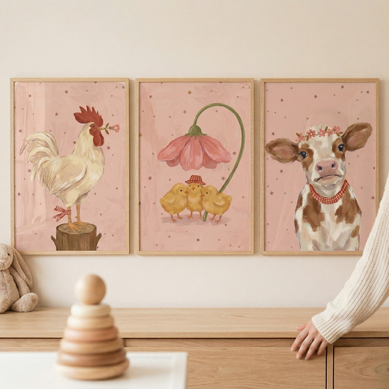 Three framed whimsical illustrations hang on a soft pink wall above a light wood dresser. The left frame shows a white rooster standing on a stump holding a flower in its beak. The center frame depicts three small yellow chicks sheltered by a large, drooping pink flower. The right frame features a brown and white cow wearing a floral crown and a checkered collar. All three have starry pink backgrounds. A wooden stacking toy and a plush rabbit are visible in the foreground.
