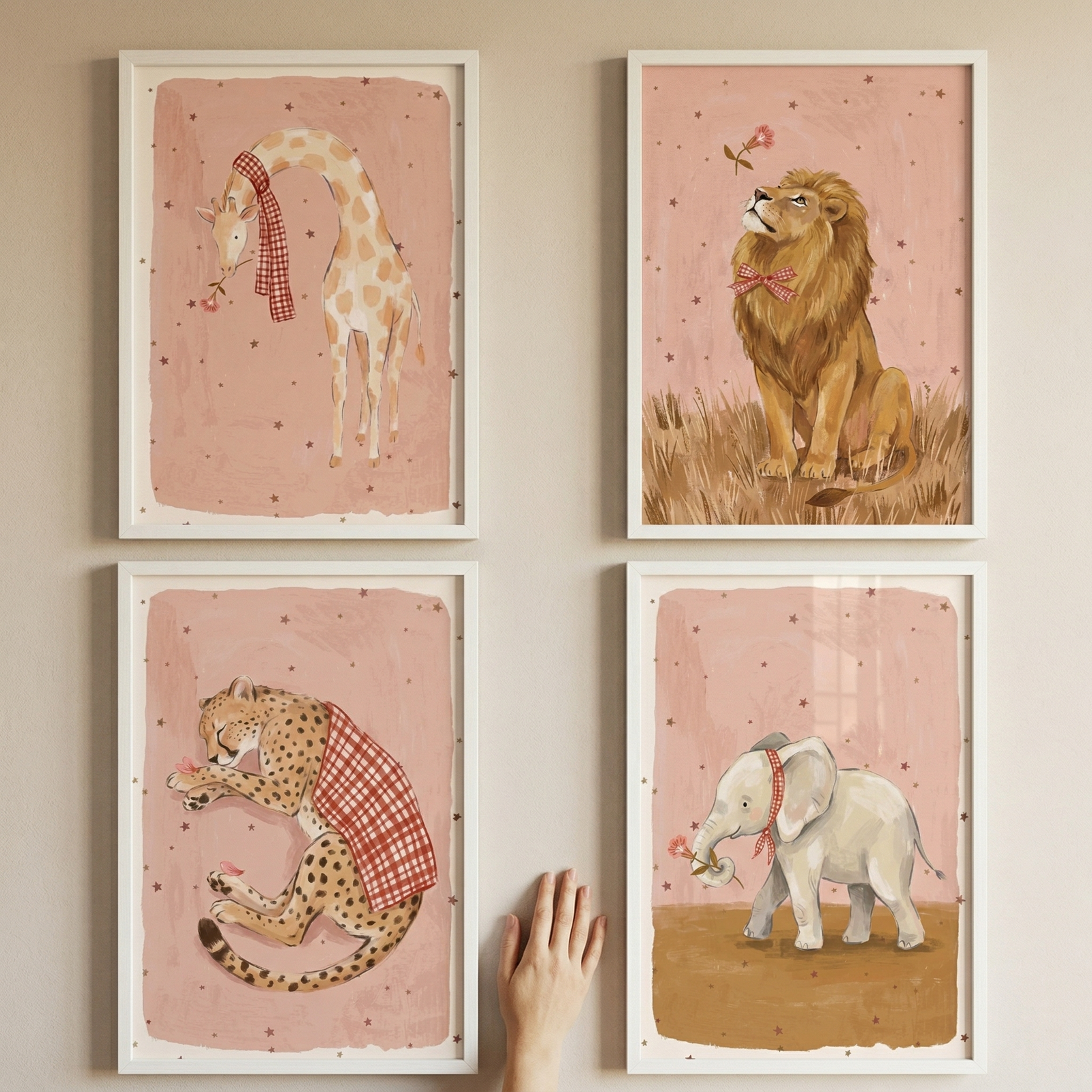 Four white-framed whimsical animal illustrations are displayed in a grid on a light beige wall. Each piece of art has a dusty pink background with tiny dark stars. The top-left frame shows a giraffe wearing a red and white checkered scarf. To its right, a lion with a red bow sits in dry grass. 