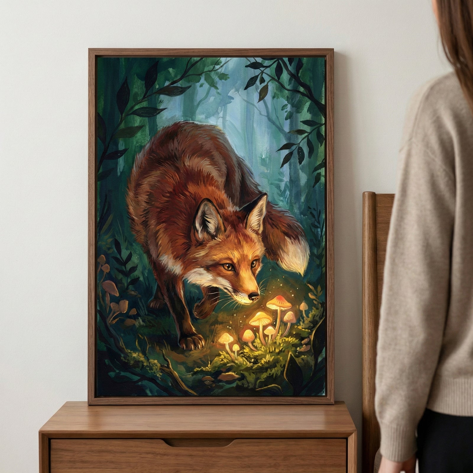 A framed piece of art showing a red fox in a dense forest, focusing on a group of small, glowing mushrooms on the mossy ground. The fox's face is illuminated by the soft light from the mushrooms. The forest background is filled with dark green leaves and tree trunks. The frame is placed on a wooden cabinet, with a person in a light-colored sweater standing nearby.