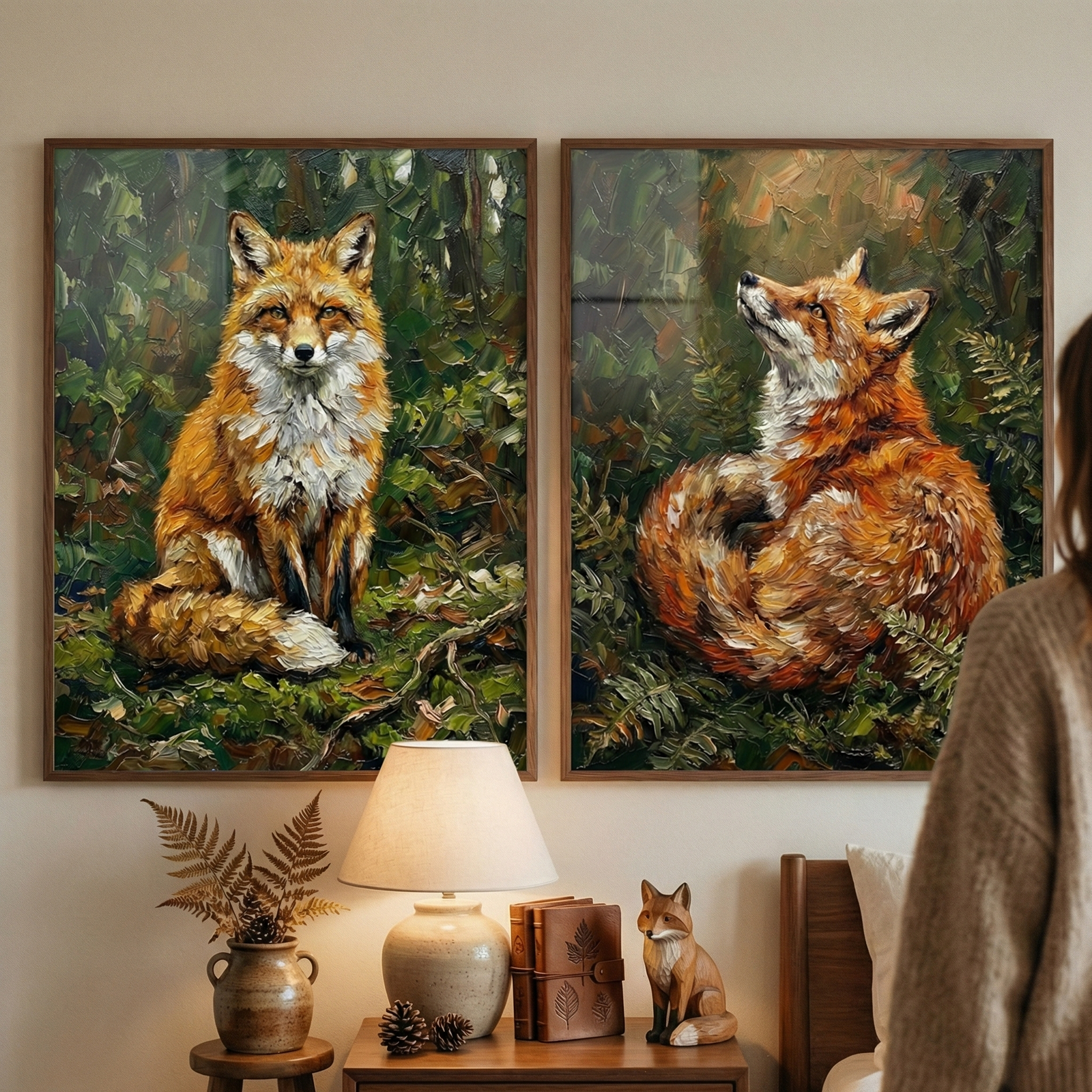 Two large, framed artworks of foxes hang on a wall above a wooden nightstand. The left piece depicts a fox sitting alertly, while the right shows a fox curled up, looking upward. Both use thick, expressive strokes in shades of rust and white against a lush green background. The nightstand below holds a warm lamp, leather-bound books, and a small fox figurine. To the left, a vase with dried ferns rests on a stool. A person in a beige sweater is partially visible, viewing the display.