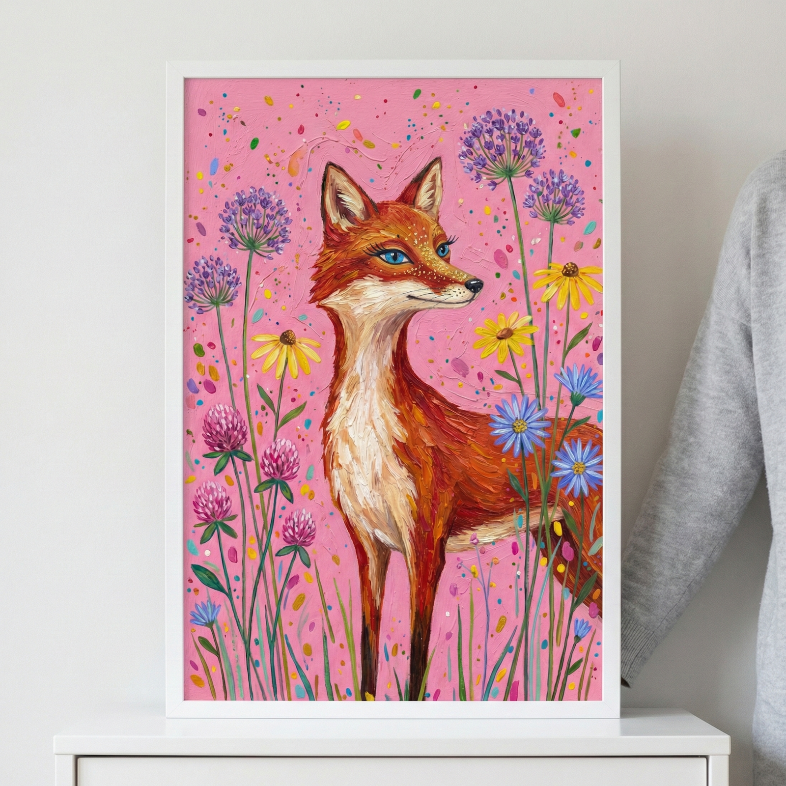A framed vertical artwork showcases a whimsical red fox standing in a vibrant meadow. The fox features thick, textured orange fur and striking blue eyes, looking gracefully to the side. It is surrounded by various colorful flowers like purple pom-poms, yellow daisies, and pink blossoms. The background is a bright pink decorated with colorful speckles. The piece is presented in a white frame, sitting on a white cabinet beside a person wearing a grey sweater.