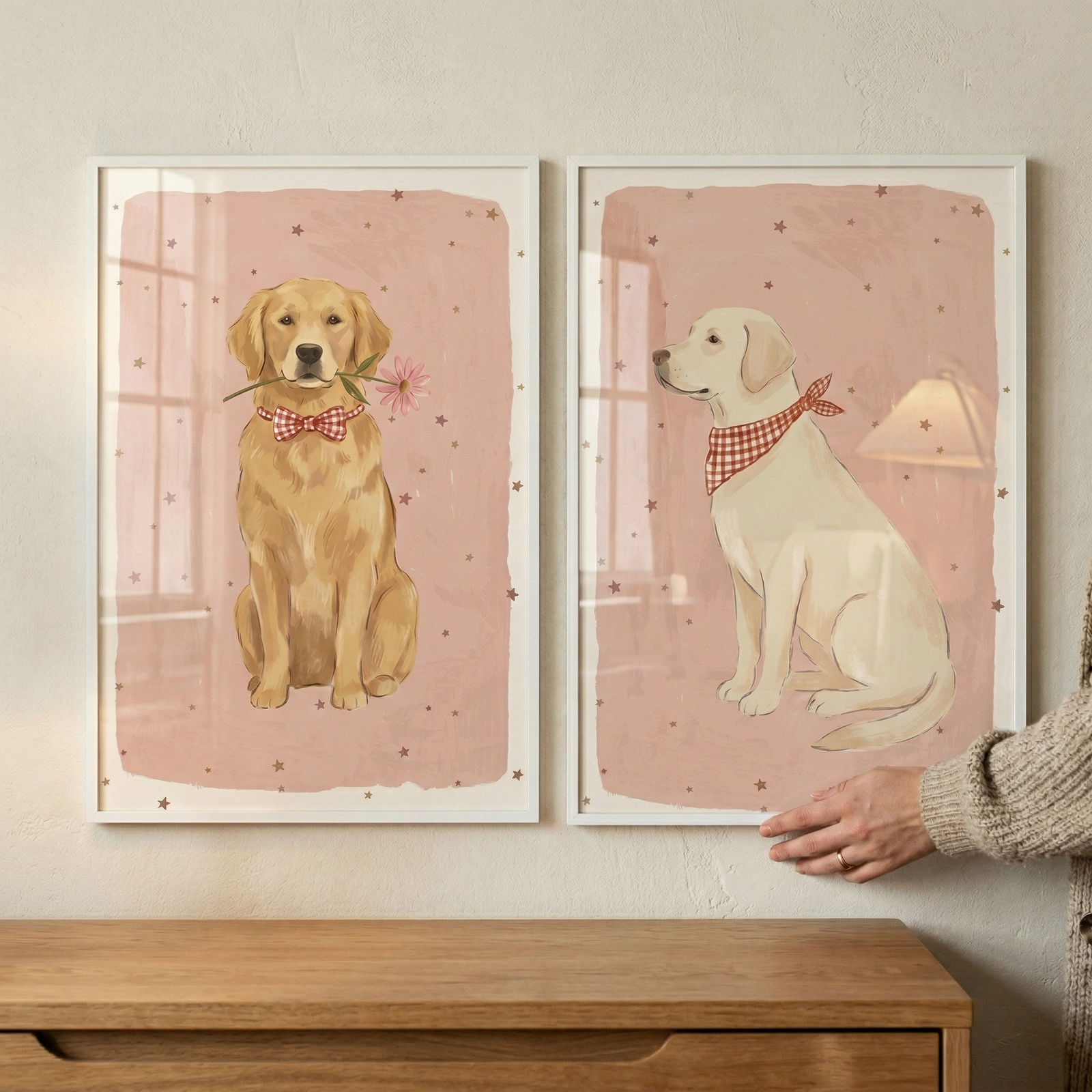 Two framed illustrations of dogs hang on a light-colored wall above a wooden chest of drawers. The frame on the left depicts a golden-furred dog sitting and facing forward, wearing a red and white checkered bow tie and holding a pink flower in its mouth. The frame on the right shows a light-colored dog sitting and facing left, wearing a red and white checkered bandana. 