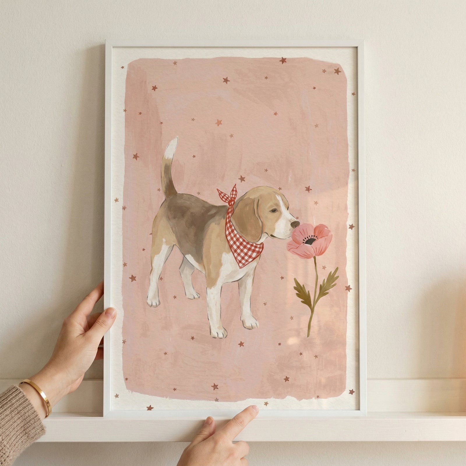 Framed illustration of a small brown and white dog wearing a red checkered bandana standing beside a pink flower on a soft blush background with scattered stars. A person’s hands hold the frame against a wall, showing the scale and adding a warm, styled nursery decor feel.