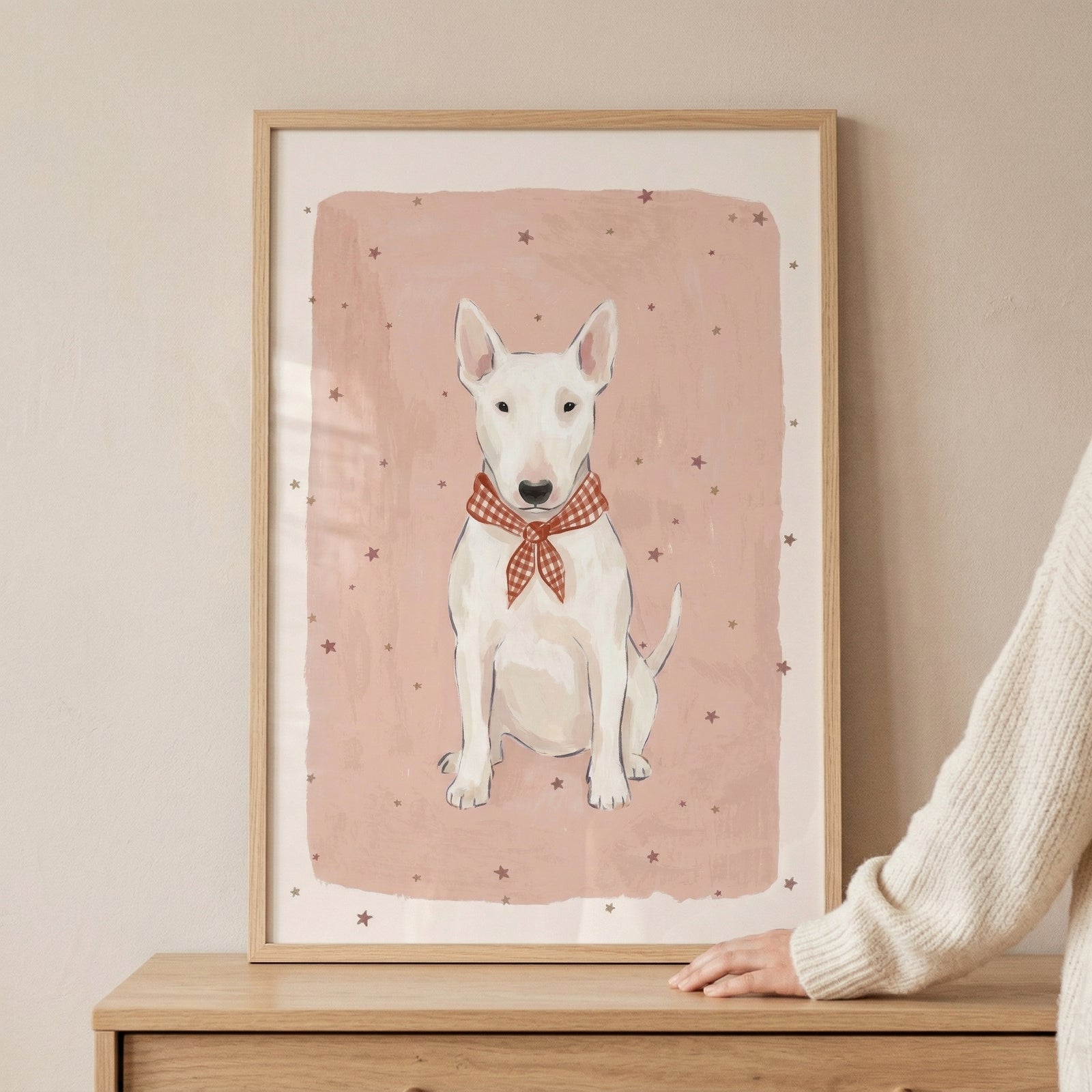 A framed illustration featuring a white Bull Terrier sitting frontally. The dog wears a red and white checkered neckerchief tied in a bow. The background of the artwork is a soft, muted pink wash with small, dark stars scattered throughout. The piece is held in a light-toned wooden frame resting on a wooden cabinet, with a person's hand in a cream knit sleeve resting against its side.