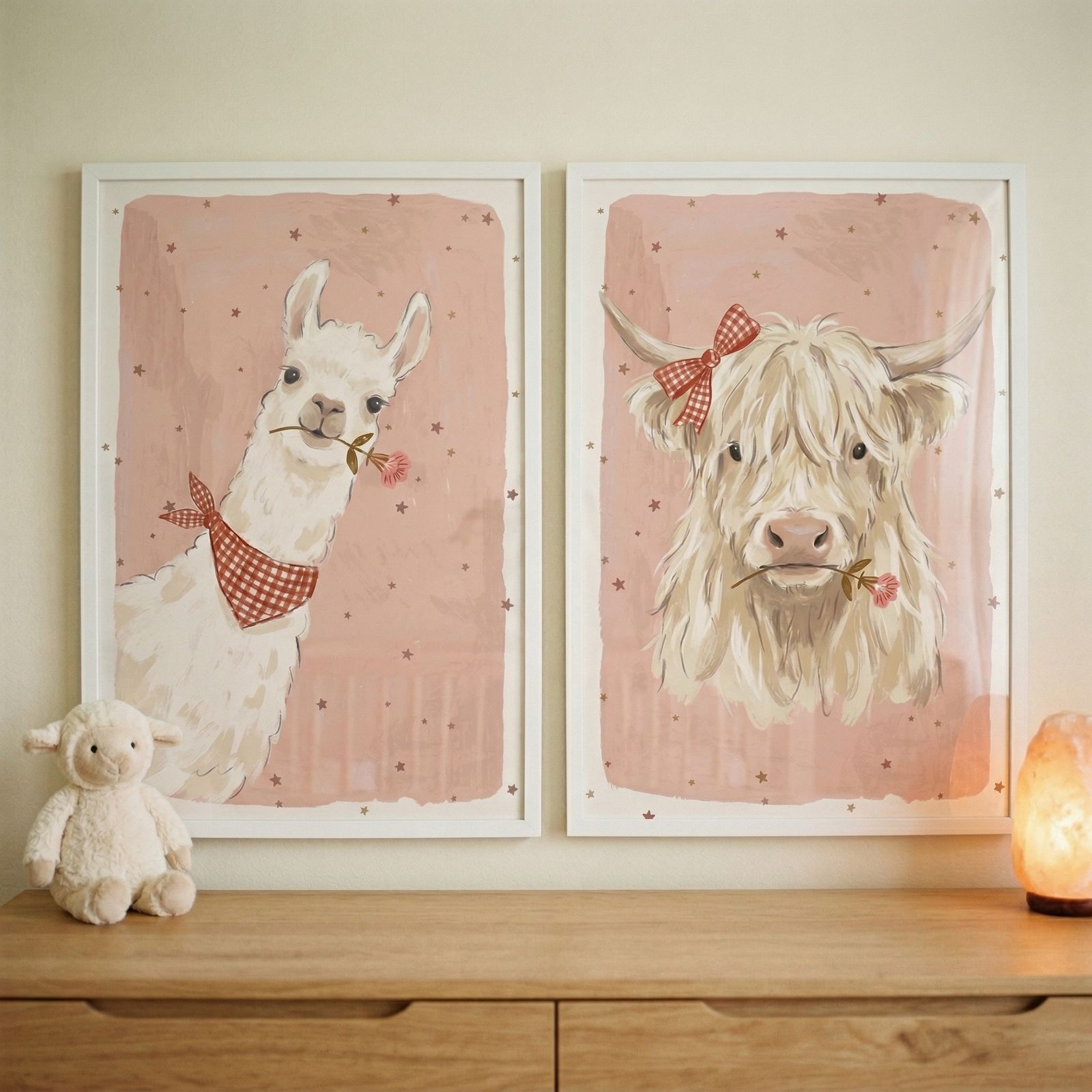Two framed illustrations hang on a neutral wall above a wooden dresser. The left frame shows a cream llama with a red-and-white checkered bandana holding a pink flower. The right frame depicts a shaggy cream cow with a matching bow and flower. Both have a soft pink background with tiny stars. A white plush sheep sits on the left of the dresser, and a glowing orange lamp is on the right, casting a warm light.