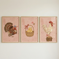 Three framed illustrations are hung side-by-side on a neutral-toned wall. Each artwork has a light wood frame and a dusty pink background with tiny dark stars. The first illustration on the left shows a brown turkey wearing a red bow and holding a pink flower in its beak. The middle illustration features a fluffy yellow chick sitting in a woven basket adorned with a red and white checkered bow. The illustration on the right depicts a white rooster on a tree stump, also holding a pink flower.