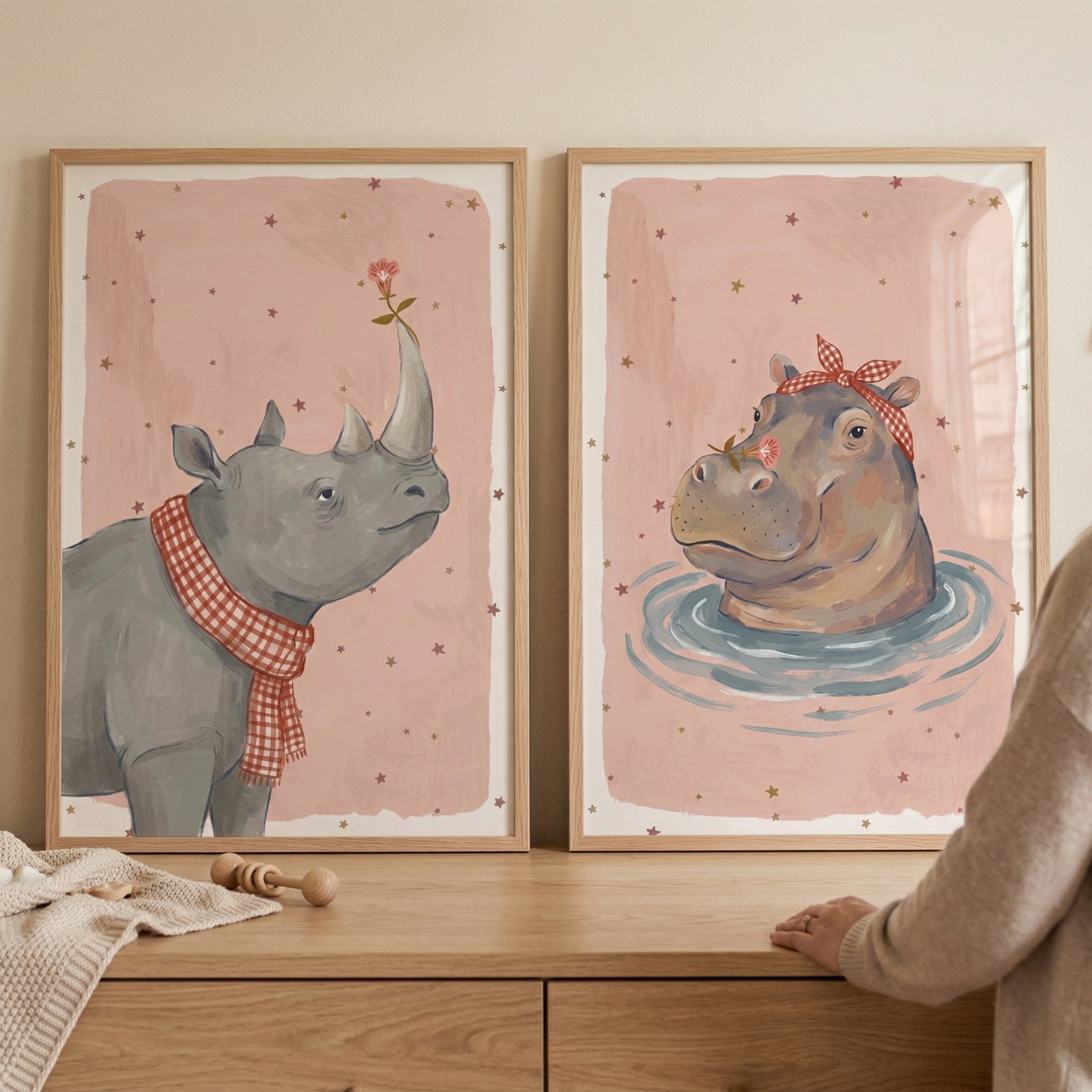 Two framed whimsical animal illustrations rest on a light wood surface. On the left, a gray rhinoceros wears a red and white checkered scarf and has a small flower on its horn. On the right, a hippopotamus peeks from blue water wearing a matching checkered headband. 