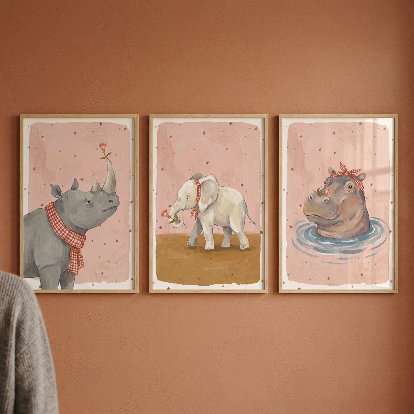 Three framed illustrations hang on a reddish-brown wall, featuring whimsical animals on soft pink backgrounds. The left piece shows a grey rhinoceros with a red checkered scarf and a small pink flower on its horn. The middle piece depicts a small white elephant with a red neck ribbon holding a pink flower in its trunk. The right piece shows a hippopotamus head in blue water ripples, wearing a red checkered headband. The illustrations share a cohesive, gentle style with light wooden frames.