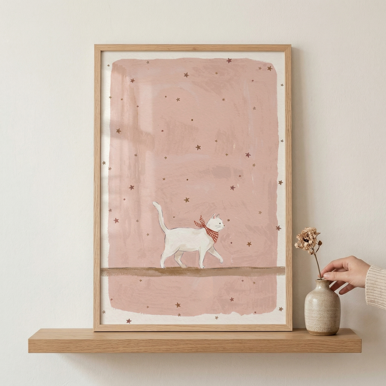 A framed illustration sits on a light wooden floating shelf against a neutral wall. The art depicts a whimsical white cat with a long tail wearing a red and white checkered scarf, walking along a brown branch. The background is a soft, textured pink wash adorned with small scattered stars. Beside the frame, a small ceramic vase holds a dried sprig, with a hand reaching out to touch the vase.