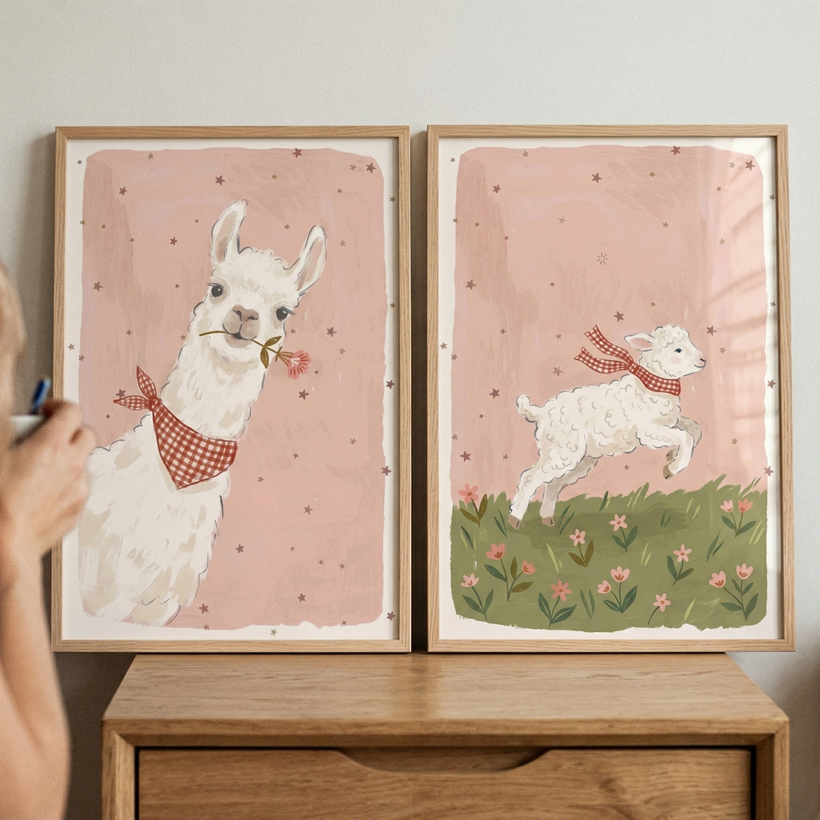 Two framed pastel animal illustrations sit on a wooden dresser showing a llama and a lamb both wearing red checkered scarves and holding flowers. A hand enters from the side adjusting the artwork, while the soft pink star patterned backgrounds and floral details create a gentle nursery style scene.