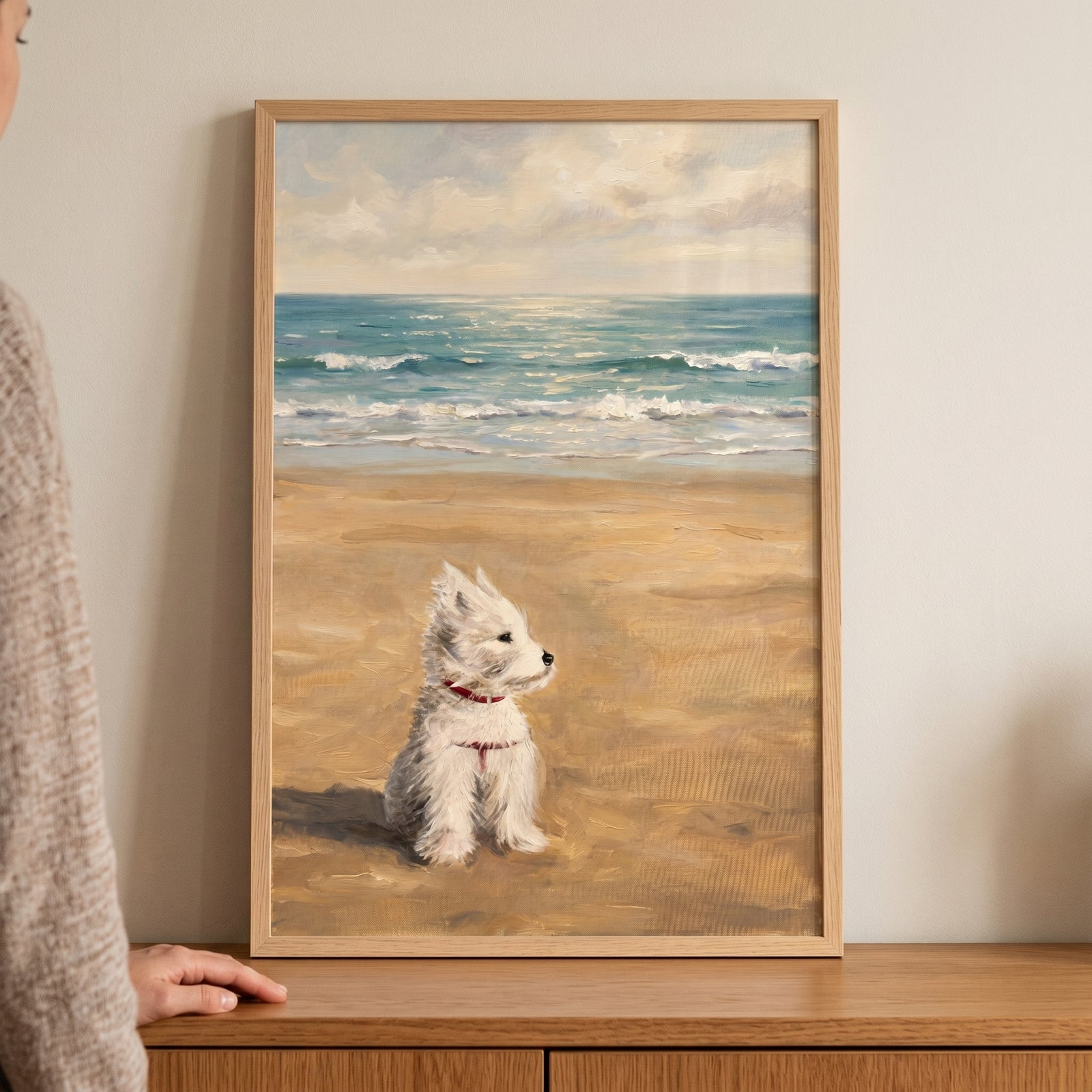 A hand sits on a wood table near framed red harness white westie dog wall art of a dog on a beach by ocean waves.