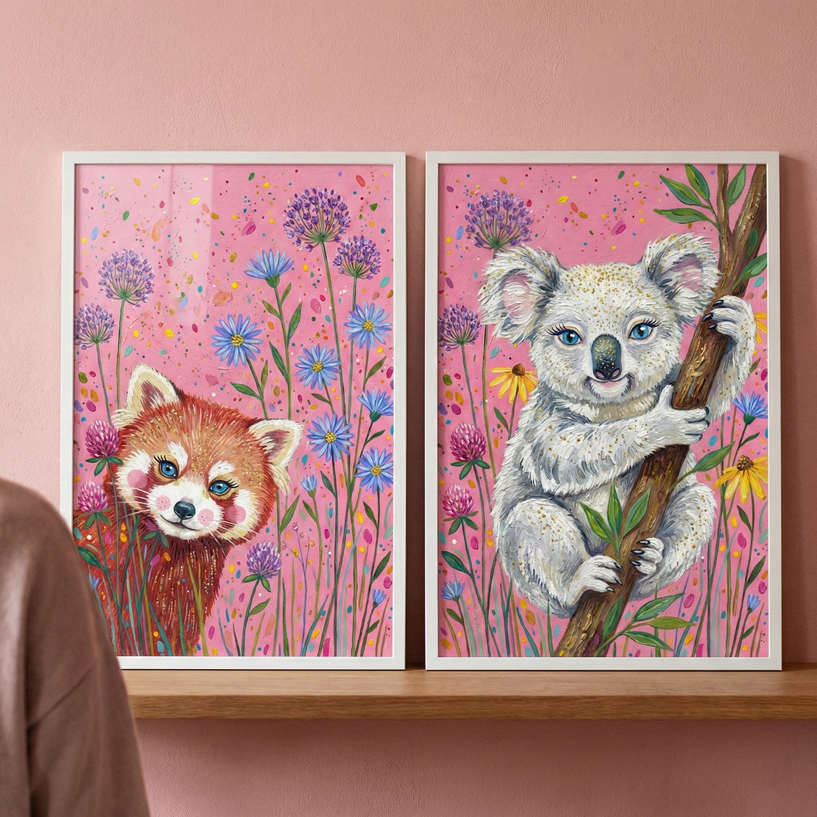 Two vertical framed artworks are displayed side-by-side on a light wooden shelf against a solid pink wall. Each piece features a stylized, whimsical animal amidst a variety of colorful flowers on a vibrant pink background. The artwork on the left depicts a red panda with fluffy reddish-brown fur, a white face, and large, expressive blue eyes. The animal is shown from the chest up, peering out from behind a collection of tall green stems and colorful blossoms. 