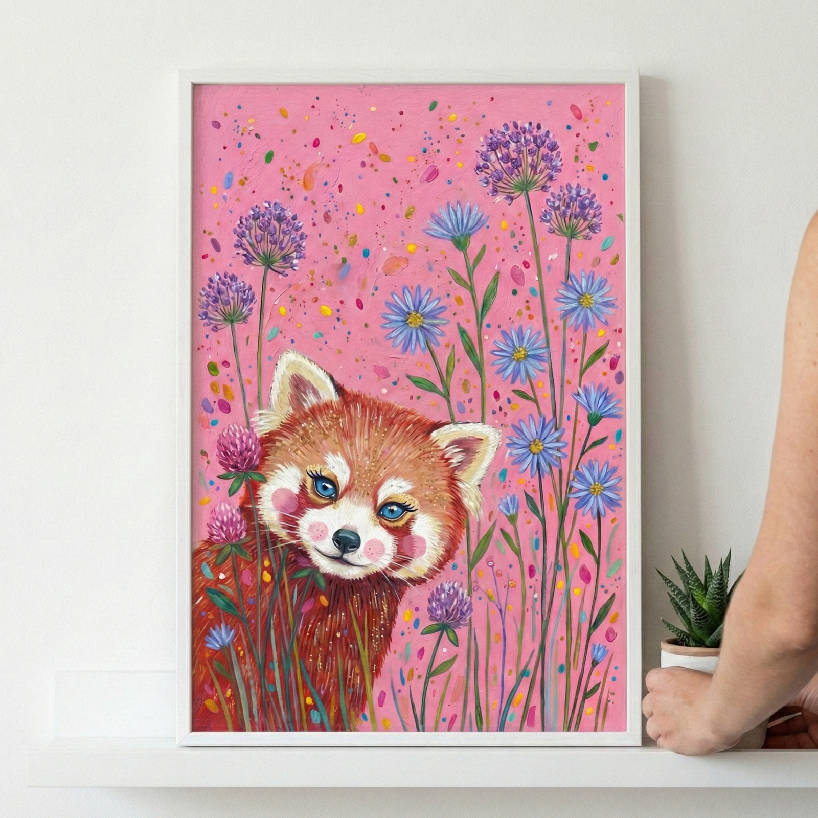 A framed illustration features a charming red panda with large blue eyes and rosy cheeks peering from behind tall blue and purple flowers. The background is a vibrant pink, adorned with colorful confetti-like speckles. The artwork is housed in a sleek white frame, resting on a white shelf against a plain wall. To the right, a hand holds a small green succulent in a white pot, adding a touch of nature to the scene.