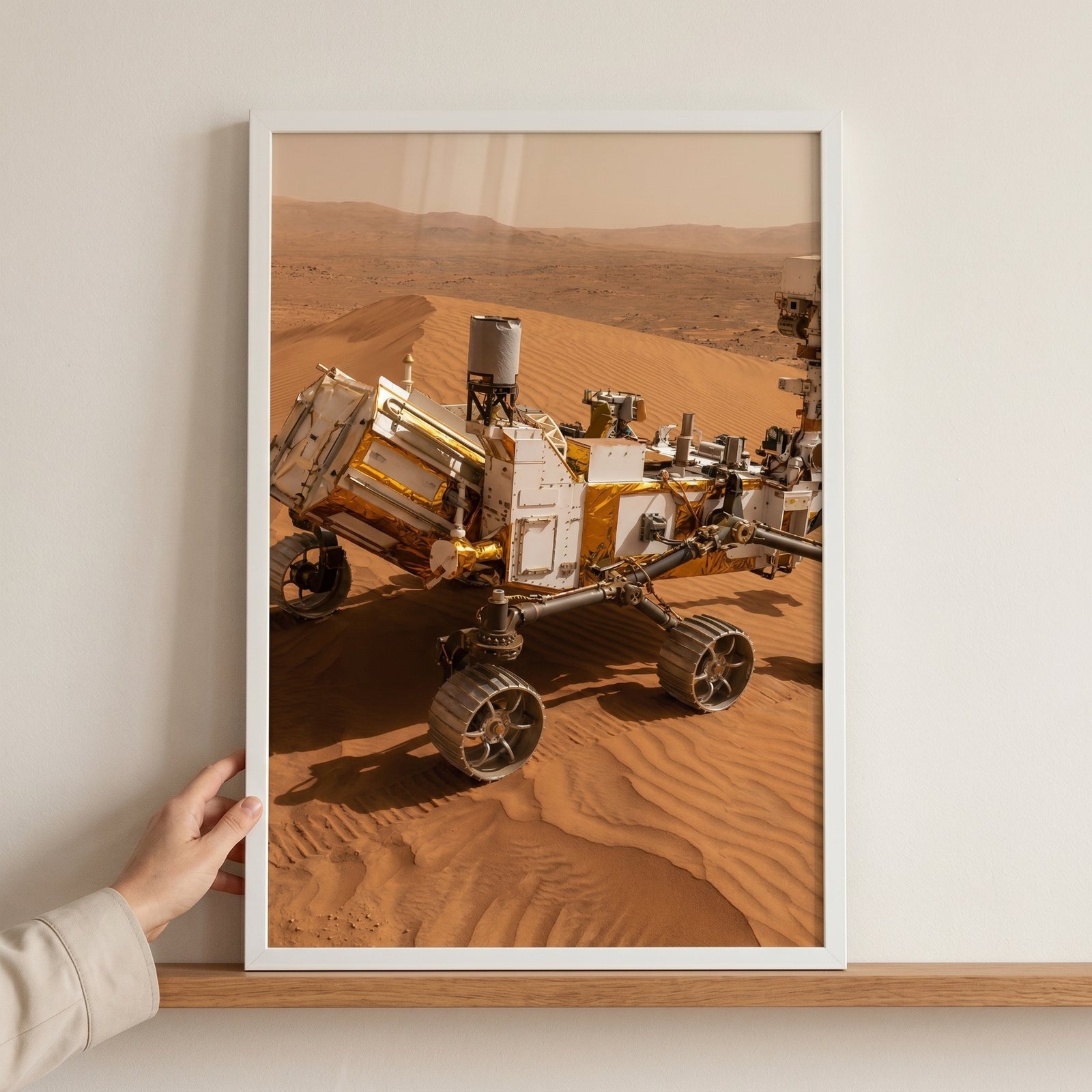 Framed red planet sand rover wall art depicts a robotic exploration vehicle on orange dunes, held by a hand on a shelf.
