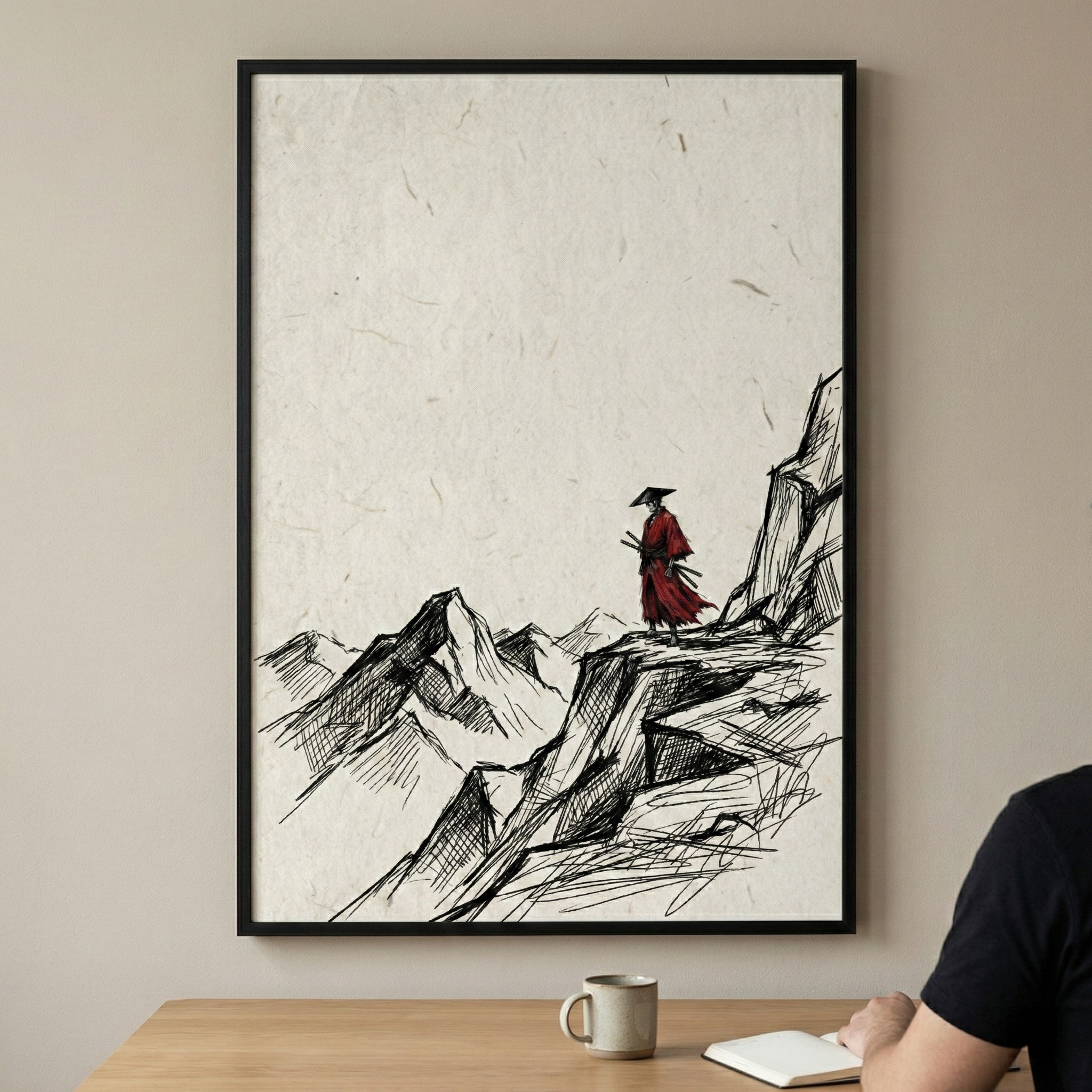 A vertical framed artwork hangs on a neutral wall above a wooden desk. The art depicts a lone figure in a bright red robe and a wide conical hat, standing at the precipice of a rocky cliff. The scene features minimalist, sketch-style mountains drawn with black ink lines on a textured off-white background. On the desk in the foreground, there is a ceramic mug and an open notebook, with a person’s arm and shoulder partially visible as they sit at the workspace.