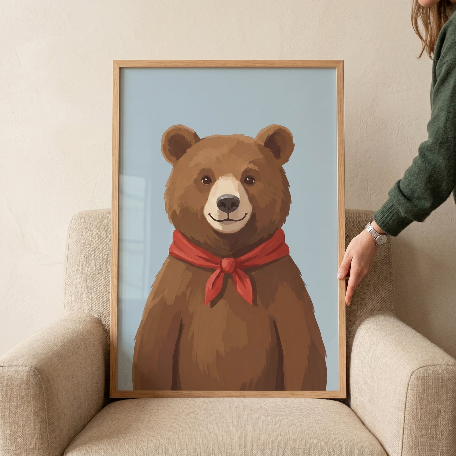 Framed red scarf brown bear wall art rests on a tan armchair held by a person's hand against a light blue background.