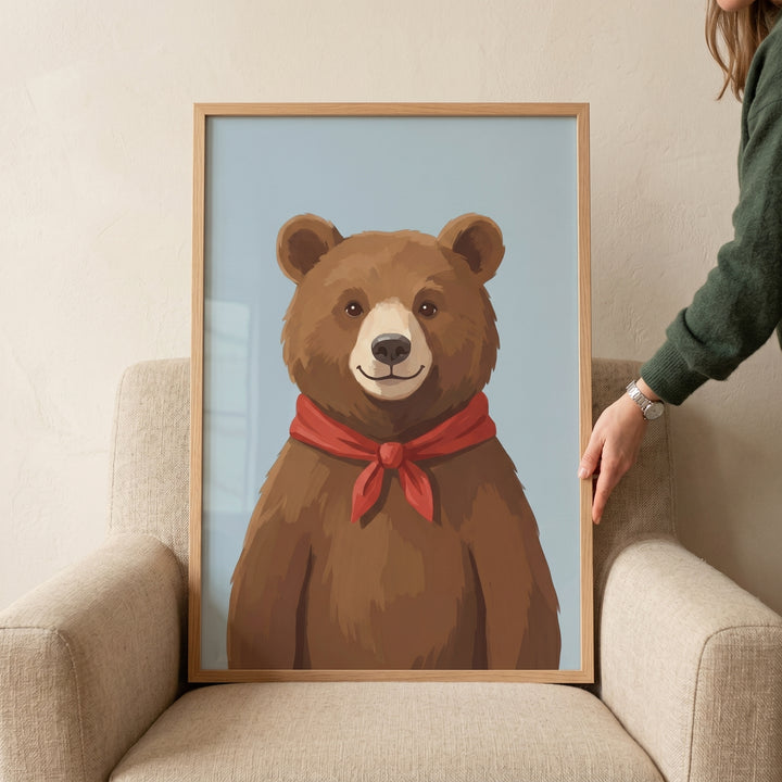Framed red scarf brown bear wall art rests on a tan armchair held by a person's hand against a light blue background.