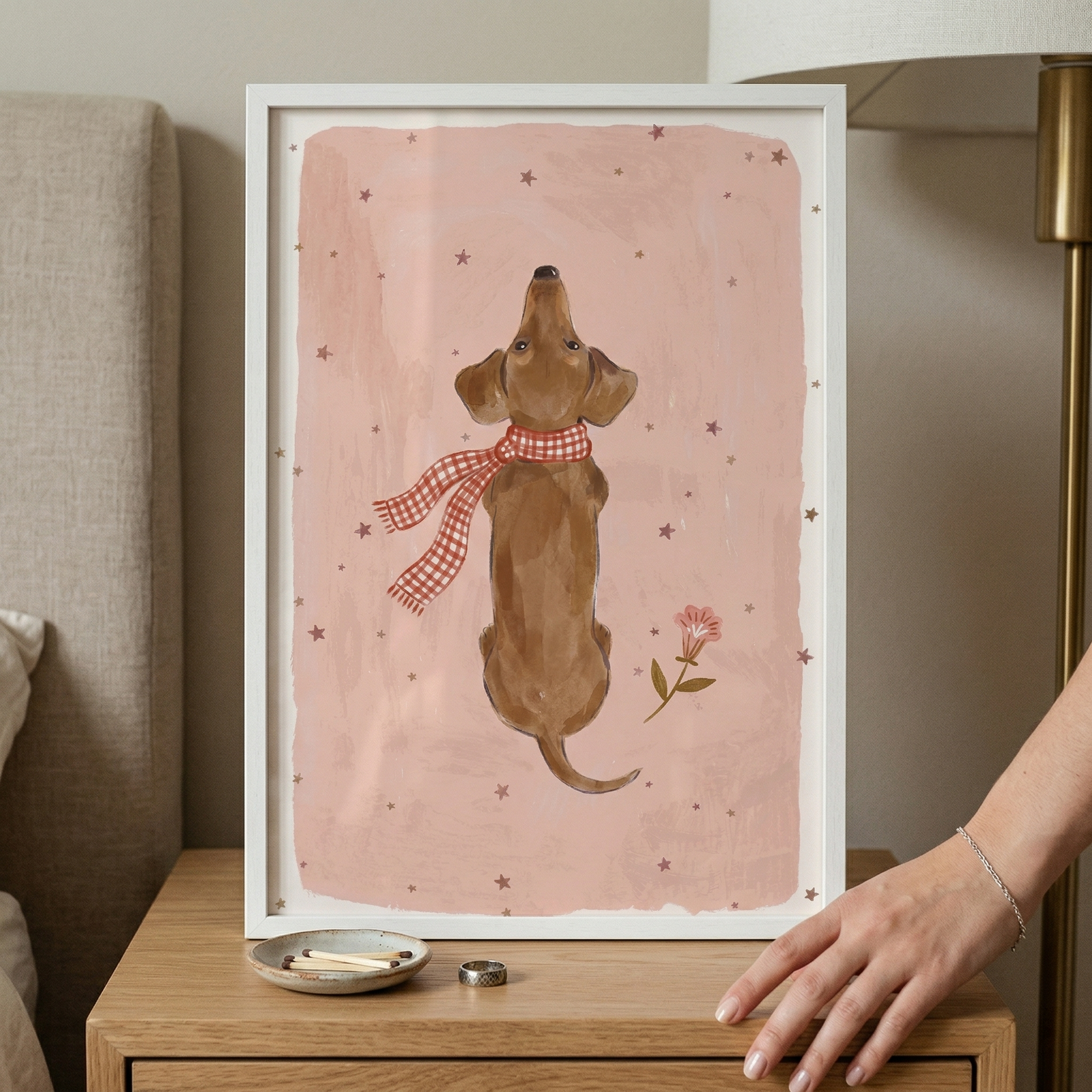 A framed illustration of a small brown dog, seen from behind as it looks up, is centered on a wooden nightstand. The dog wears a red and white gingham scarf and is set against a light pink, textured backdrop decorated with small, scattered stars and a single pink flower. The white-framed piece sits next to a small dish with matches and a silver-toned ring. A gold-toned lamp is on the right, and a grey upholstered headboard is on the left. A hand with a silver bracelet reaches toward the frame.