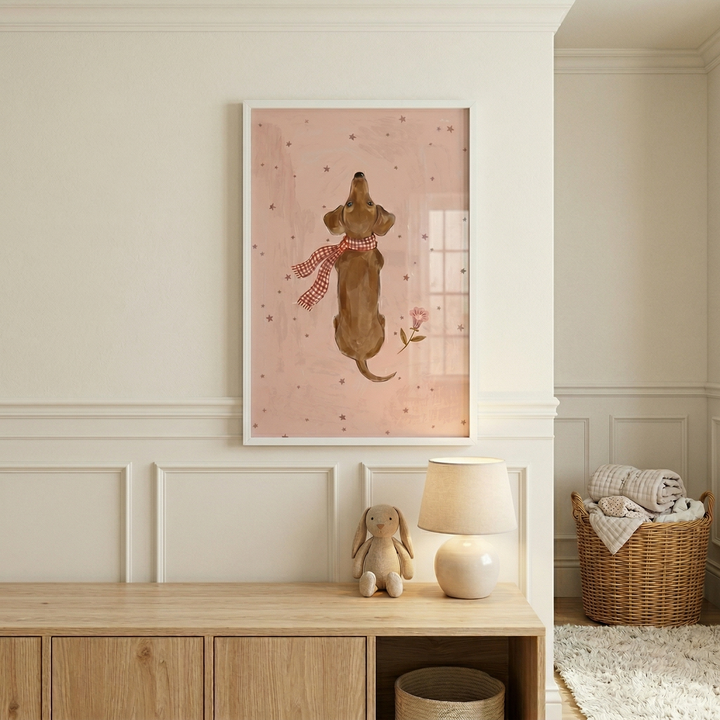A vertical framed illustration hanging on a cream wall above a light wood cabinet. The artwork depicts a brown, long-bodied dog from behind, looking upwards, wearing a red and white checkered scarf. The dog is centered on a dusty pink background with small dark stars and a tiny pink flower. On the cabinet sits a small stuffed rabbit toy and a table lamp with a round white base. To the side, a tall wicker basket is filled with folded blankets, resting on a white shag rug.