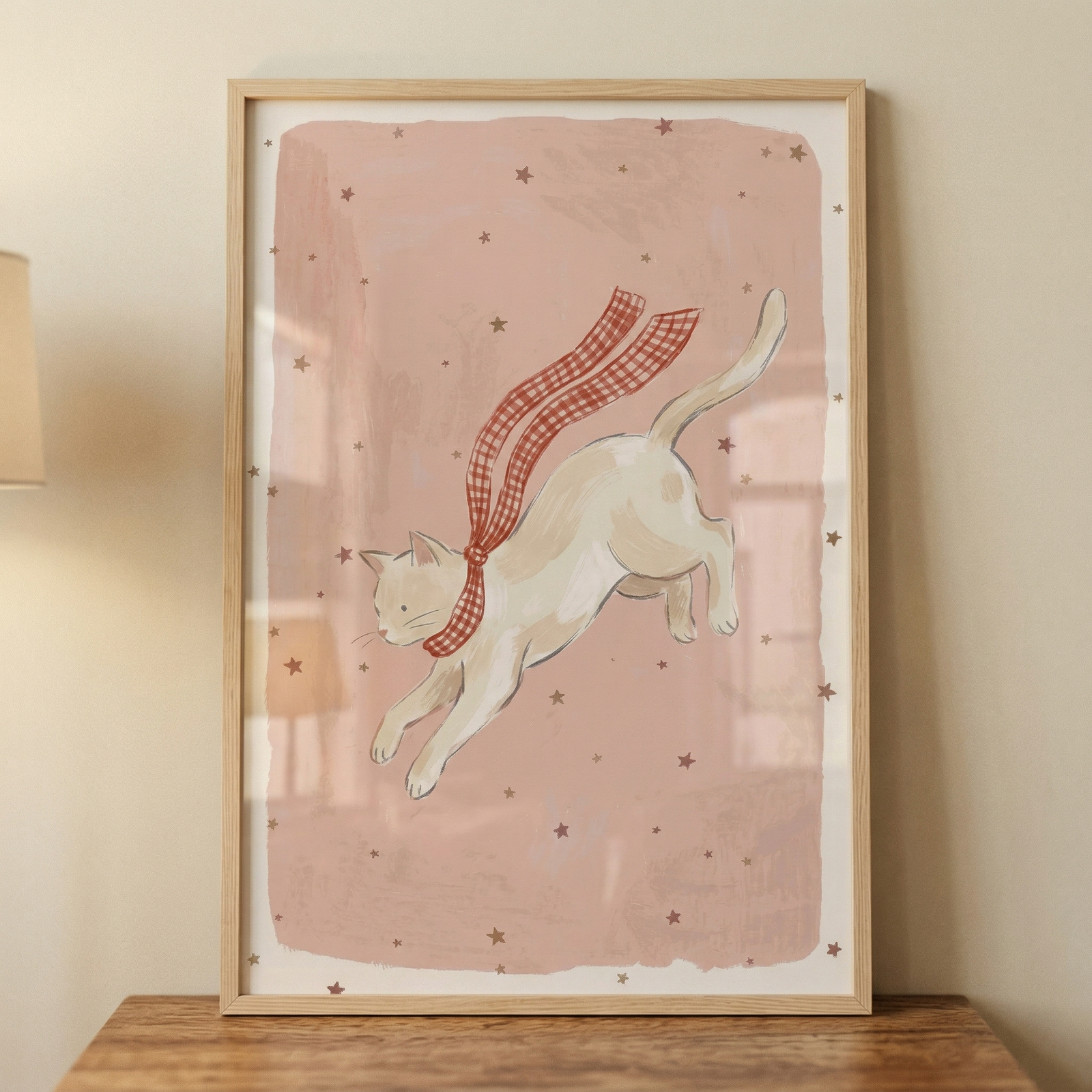 A framed illustration of a light-colored cat leaping through the air, wearing a long red and white checkered scarf that trails behind it. The cat is centered on a muted pink, textured background decorated with small scattered stars. The artwork is bordered by a white margin and housed in a light wood frame. The frame sits atop a wooden surface against a neutral-toned wall, with a soft lamp light partially visible on the left.