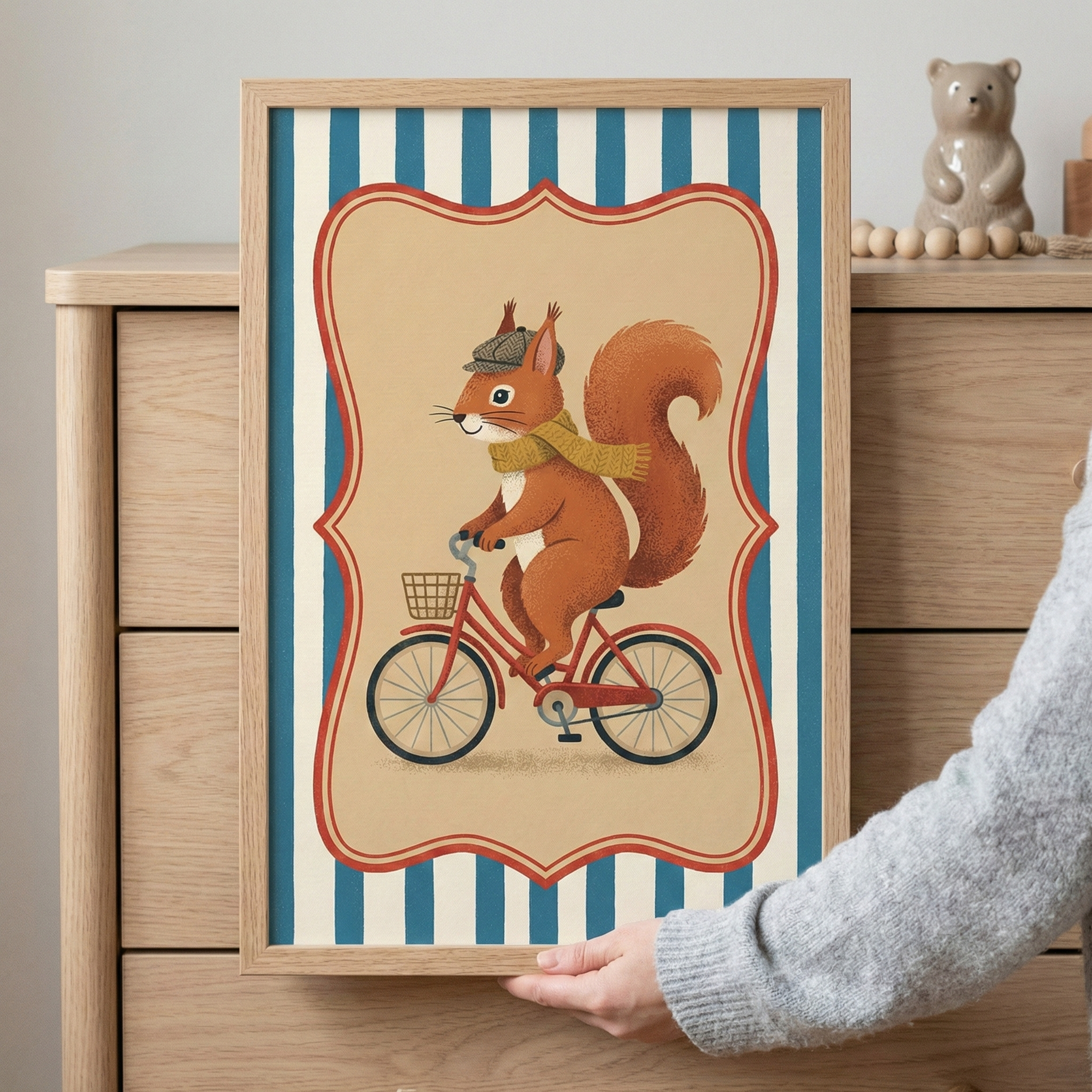 A vertical framed illustrative artwork held by a hand in front of a wooden dresser. The central illustration depicts a charming red squirrel wearing a grey newsboy cap and a yellow scarf while riding a red bicycle. The bicycle features a small woven basket on its handlebars. This central scene is contained within a decorative red-outlined scalloped frame against a background of vertical blue and cream stripes. In the soft-focus background, a small beige bear figurine and wooden beads sit on top of the dress